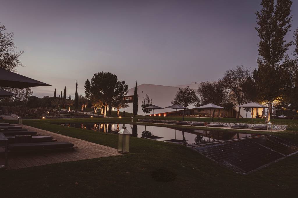 Imagem mostra o edifício principal do hotel, um caixote branco de linhas retas, com uma piscina em frente e um jardim recheado de árvores