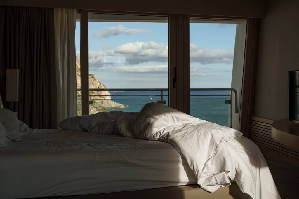 _1NC8724 Cama desarrumada num quarto de hotel, com uma varanda de vidro que revela o mar la fora