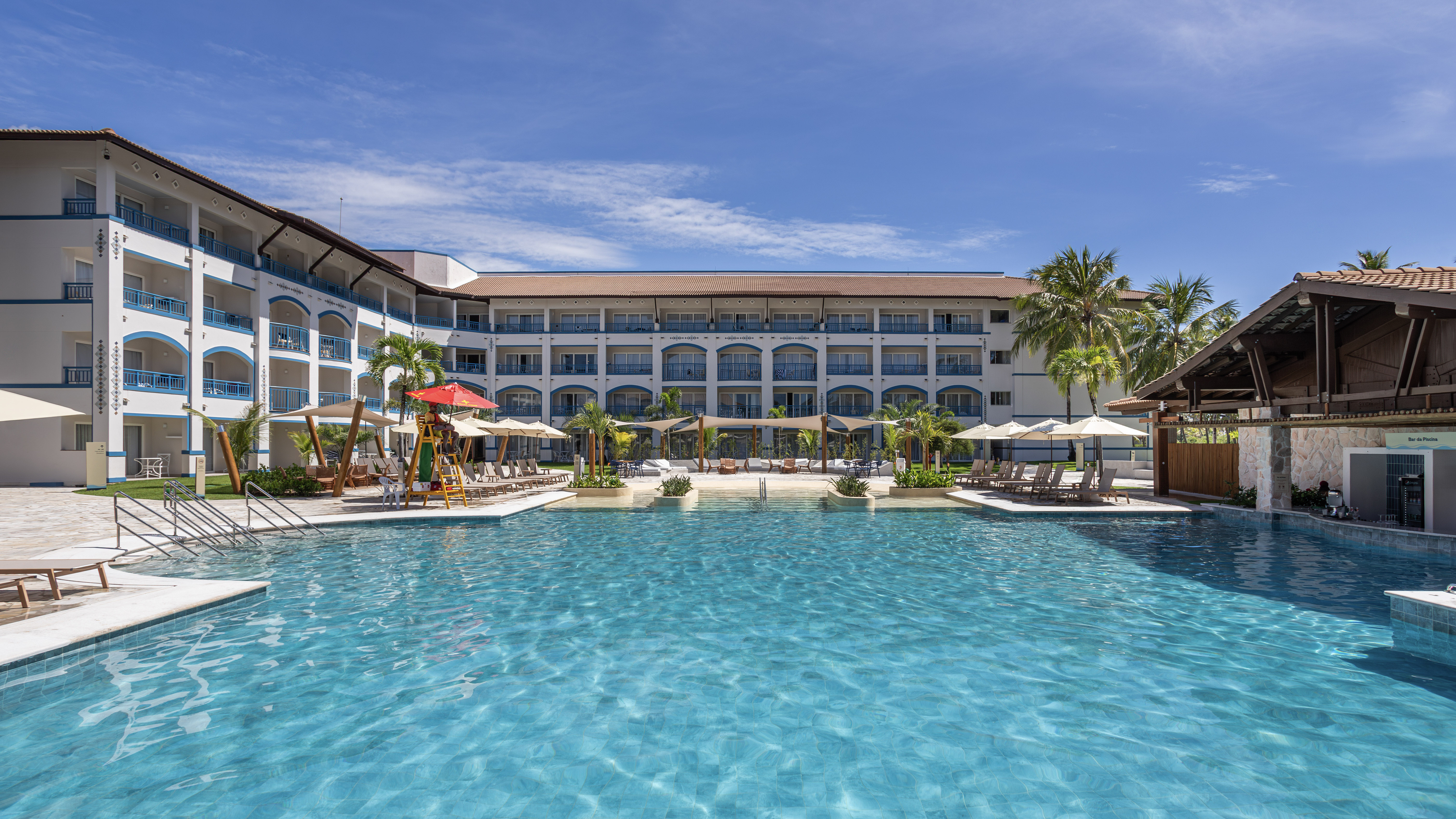 Grande piscina e, ao fundo, hotel com estilo praiano, de paredes brancas e detalhes em azul. Há guarda-sóis no entorno da piscina.