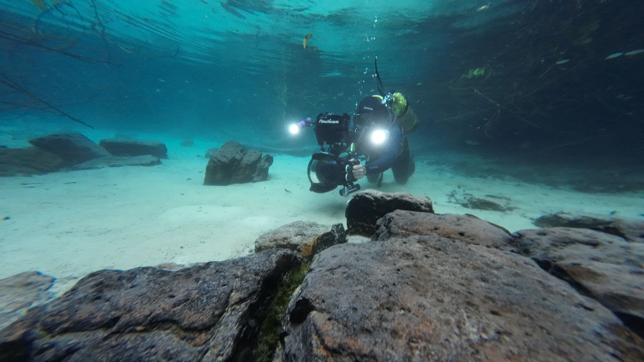 Mergulhador com equipamento de filmagem subaquática, incluindo câmera e luzes potentes, flutuando sobre um leito de areia clara com rochas escuras no fundo de um rio ou lago de águas cristalinas e azul-turquesa