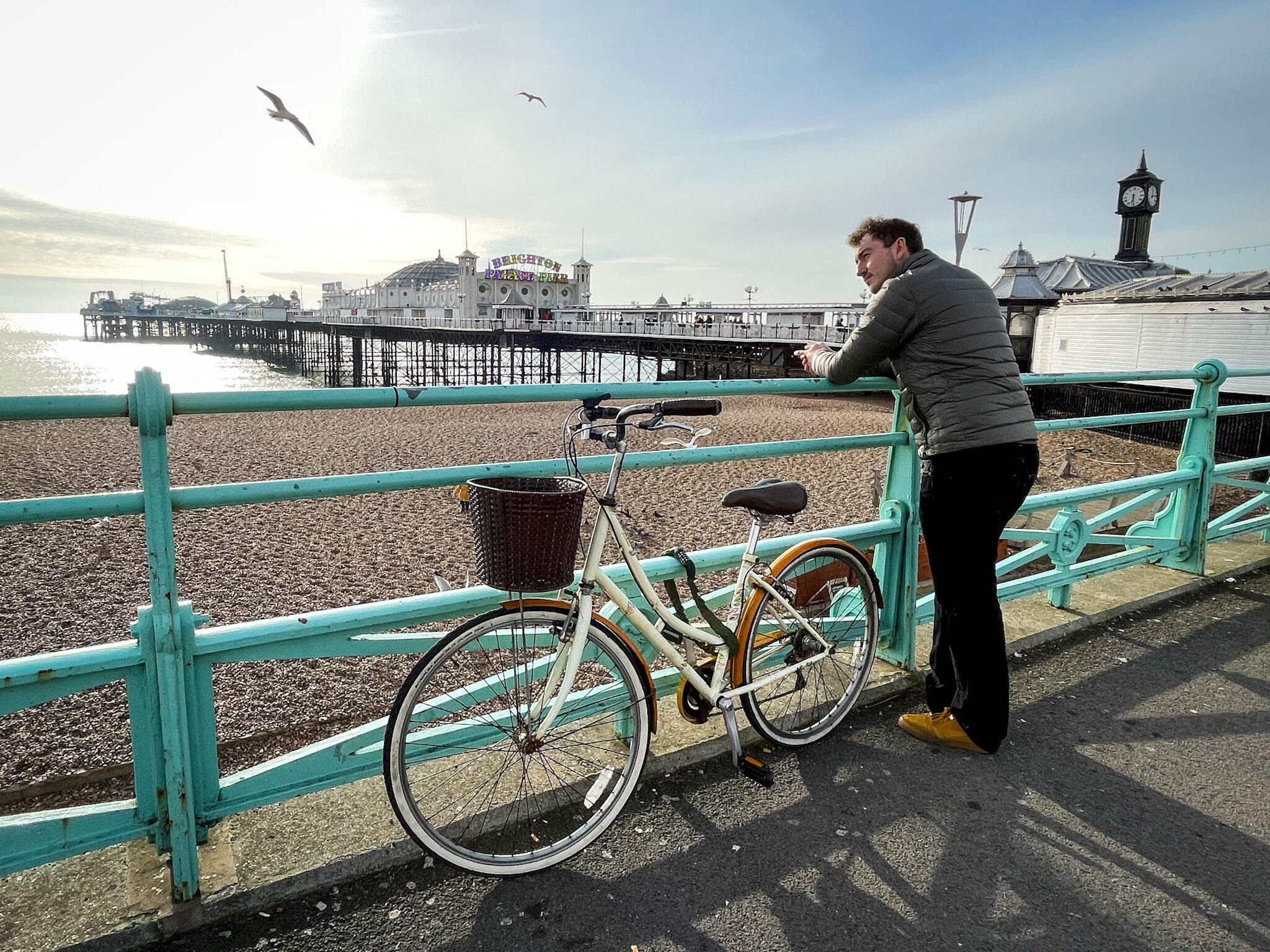 Homem de jaqueta cinza e calça preta, apoiado em uma grade verde-água, observa o mar e o píer de Brighton ao fundo. Uma bicicleta creme com cesta marrom está estacionada ao lado dele. O céu está claro, com gaivotas voando e a praia de pedras visível.
