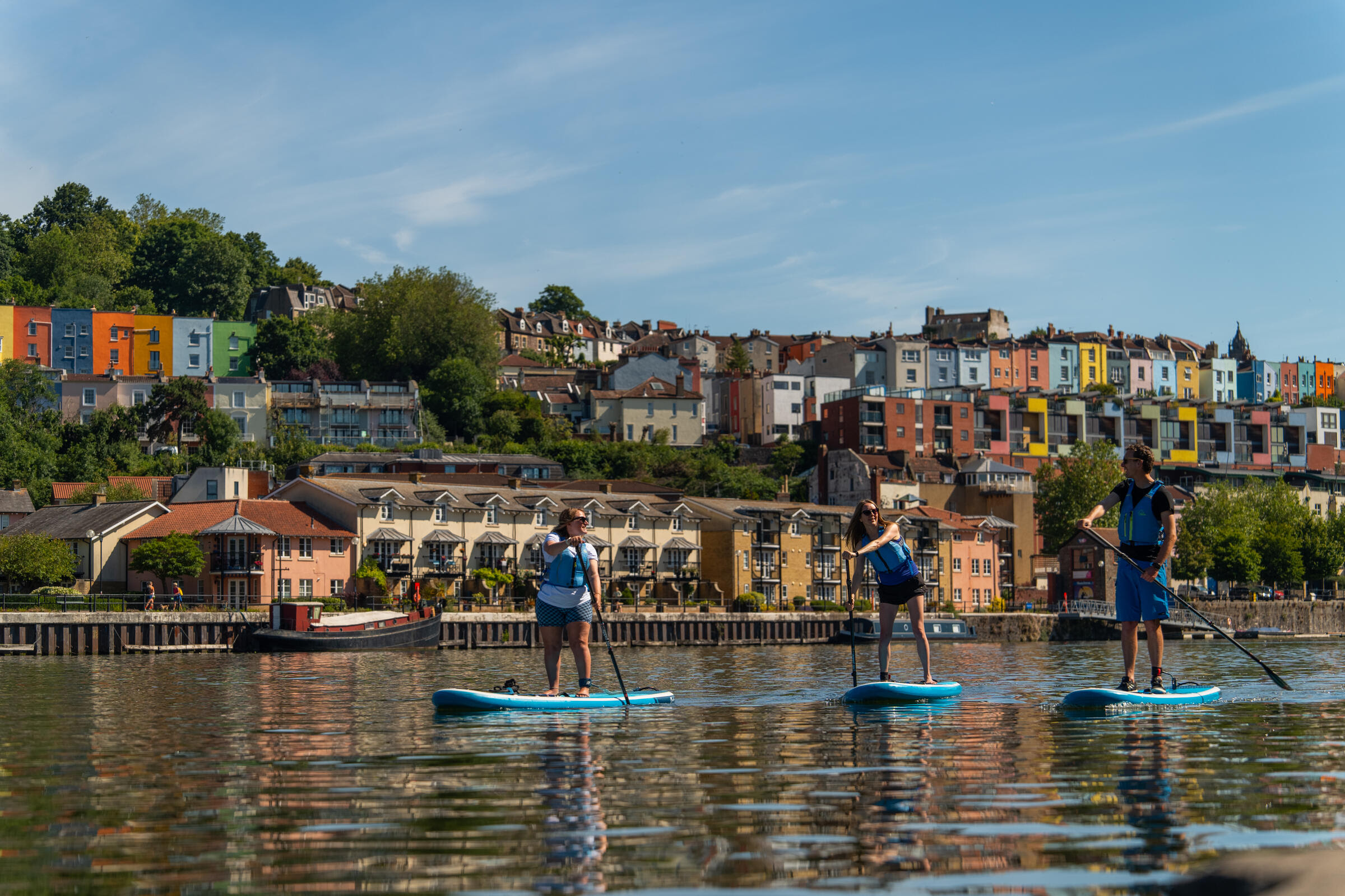 Três pessoas em pranchas de stand-up paddle remam em um rio, com casas coloridas e árvores na margem ao fundo, sob céu azul