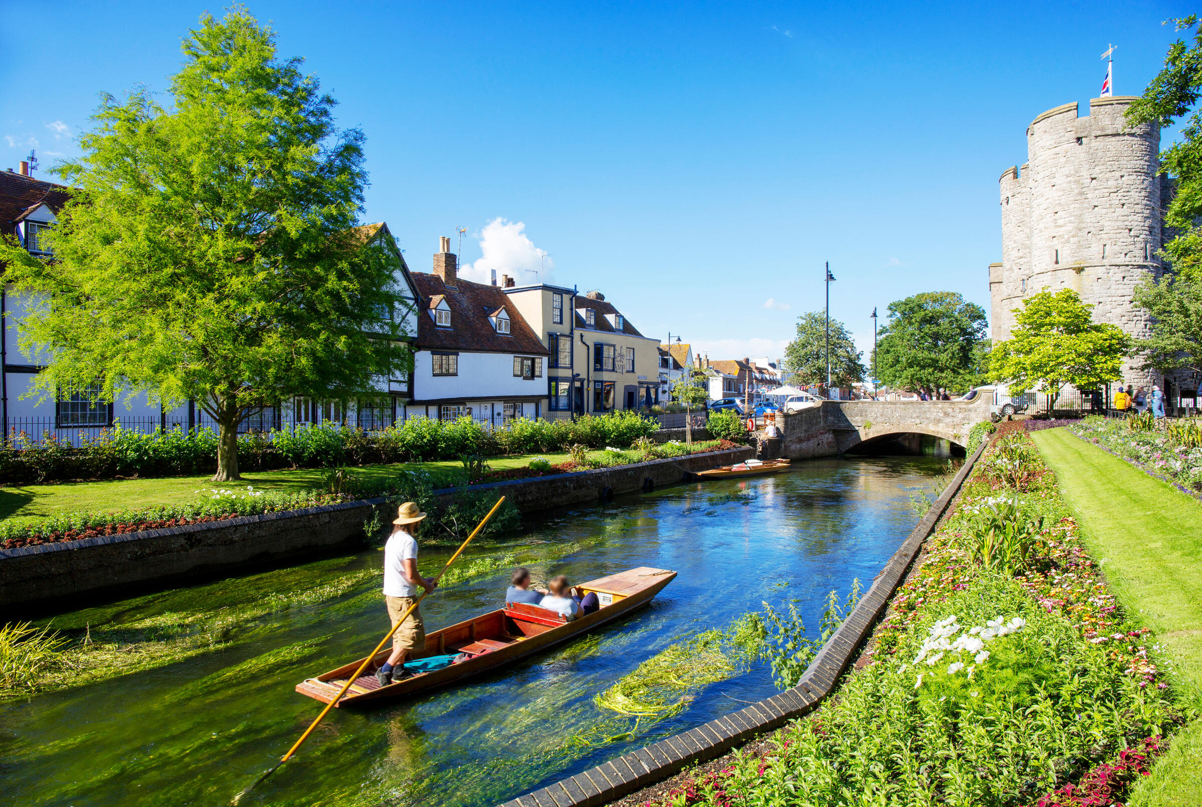 Um homem de chapéu de palha e camiseta branca empurra um barco com duas pessoas por um rio verde-azulado, ladeado por casas antigas, jardins floridos e um castelo medieval sob céu azul