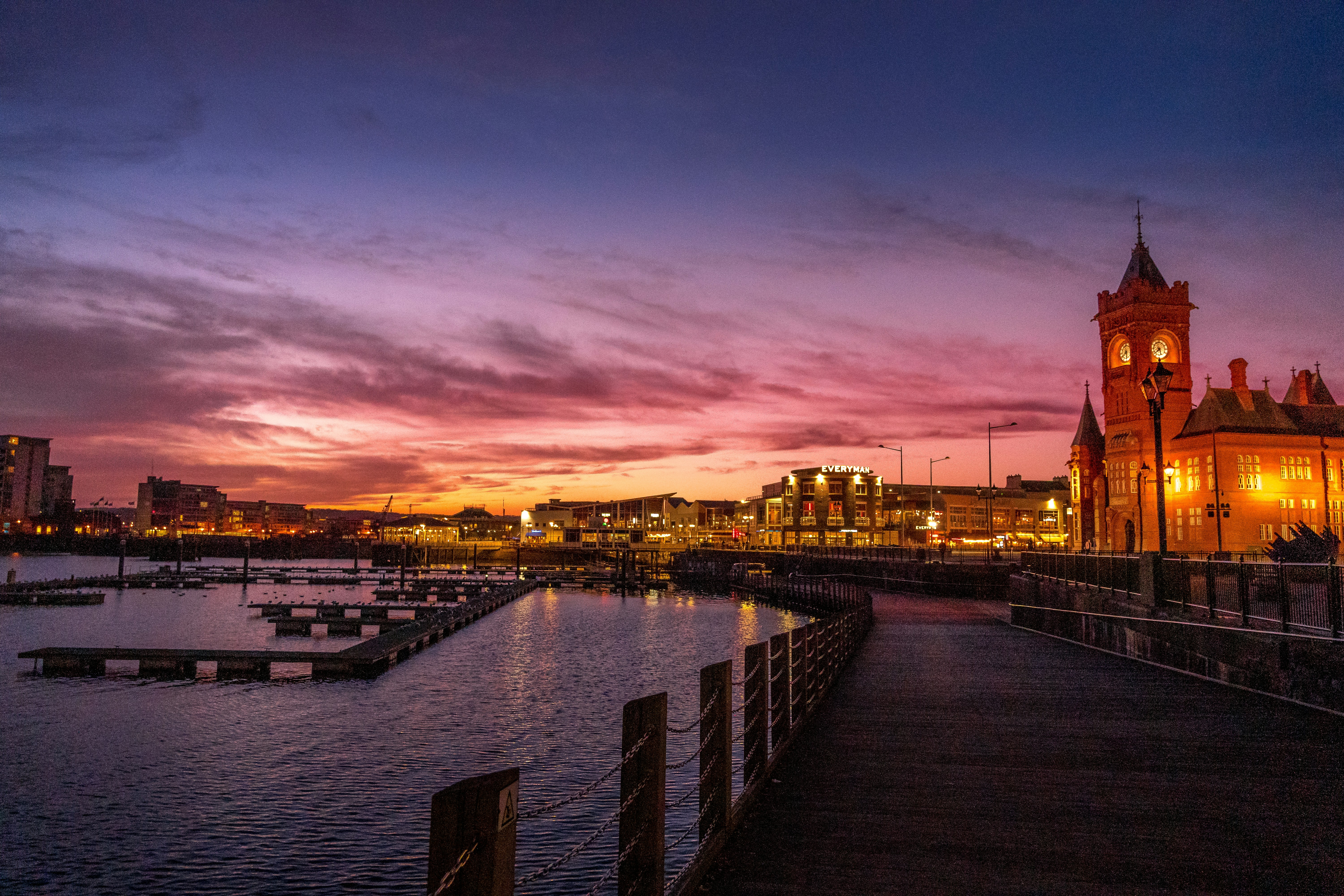 Marina com píeres vazios sob um céu de pôr do sol vibrante em tons de roxo e rosa. À direita, um edifício histórico iluminado com torre de relógio. Ao fundo, prédios modernos com luzes acesas refletem na água escura