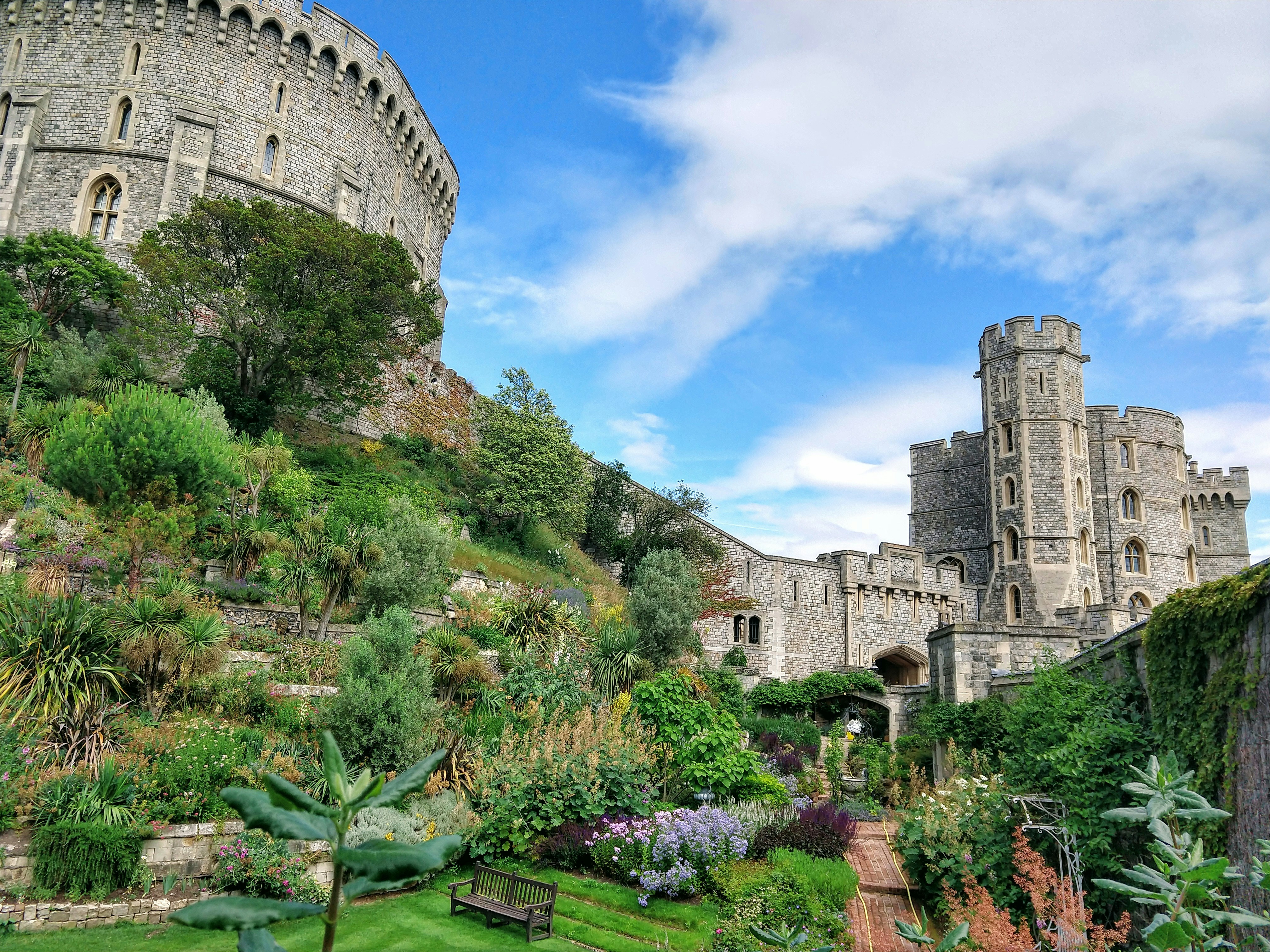 Castelo de Windsor em dia ensolarado, com torre circular à esquerda e outra torre com ameias à direita, ambas de pedra cinza. Um jardim exuberante com árvores, arbustos e flores coloridas preenche o centro e a parte inferior, com um banco de madeira no gramado e um caminho de tijolos. O céu azul tem nuvens brancas esparsas