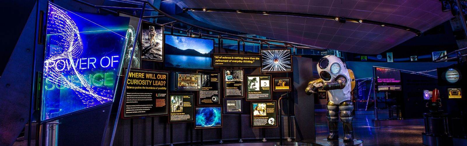 Exposição de ciência com um manequim de escafandro branco e dourado, rodeado por telas que exibem imagens de paisagens, espaço e textos informativos sobre o poder da ciência