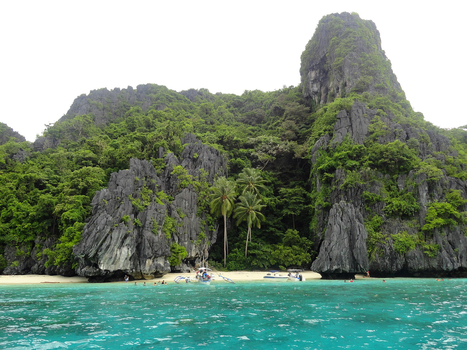 Ilha tropical com falésias rochosas escuras cobertas por vegetação densa, areia clara e mar azul-turquesa. Pequenas embarcações e pessoas nadando na água cristalina, sob céu claro