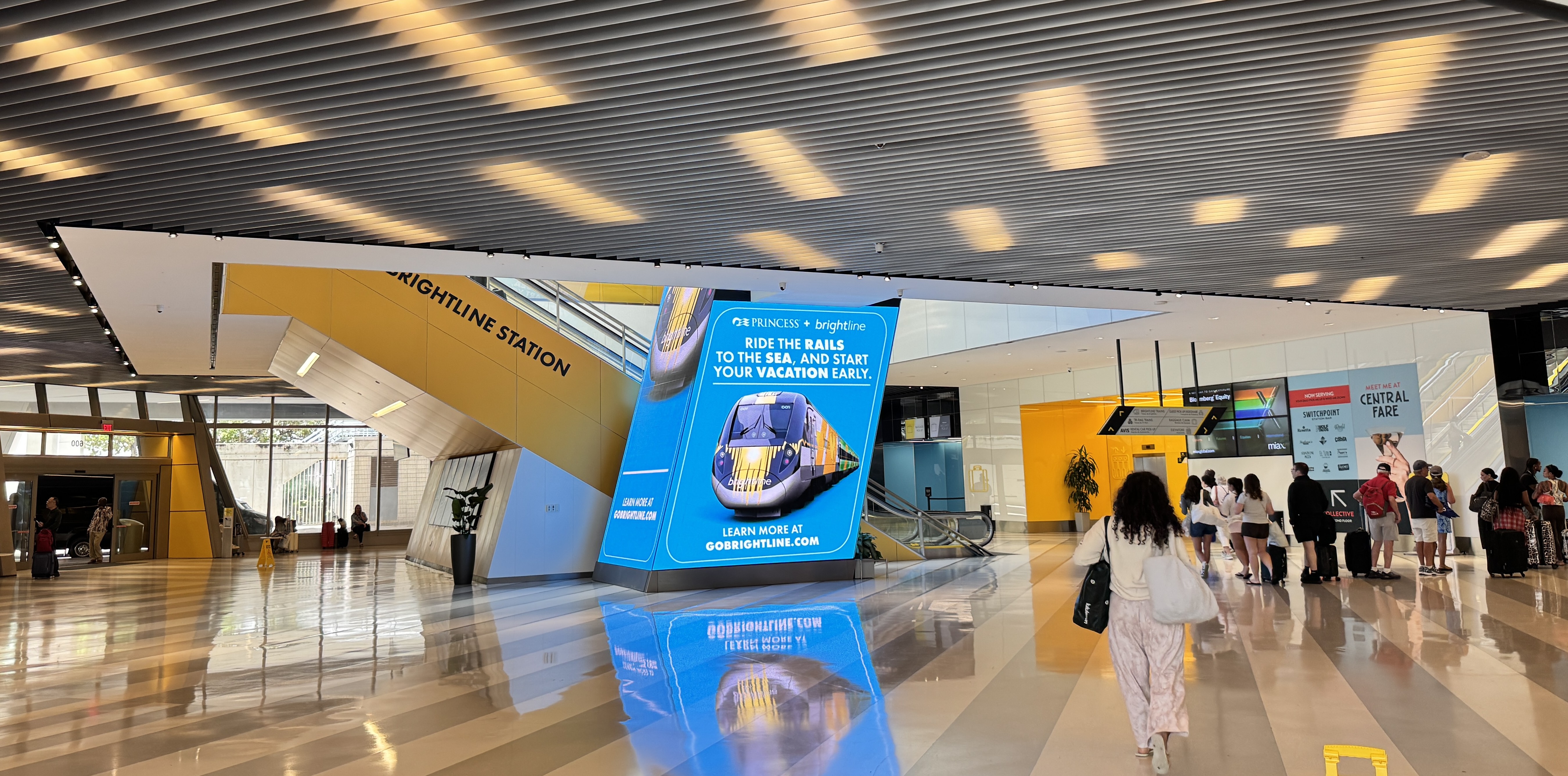 Interior de uma estação de trem moderna com teto cinza e iluminação retangular. Uma mulher de costas, com blusa clara e calça estampada, caminha em piso brilhante. À esquerda, uma estrutura amarela com BRIGHTLINE STATION e um painel azul com um trem e a frase RIDE THE RAILS TO THE SEA, AND START YOUR VACATION EARLY. Ao fundo, pessoas e balcões de atendimento