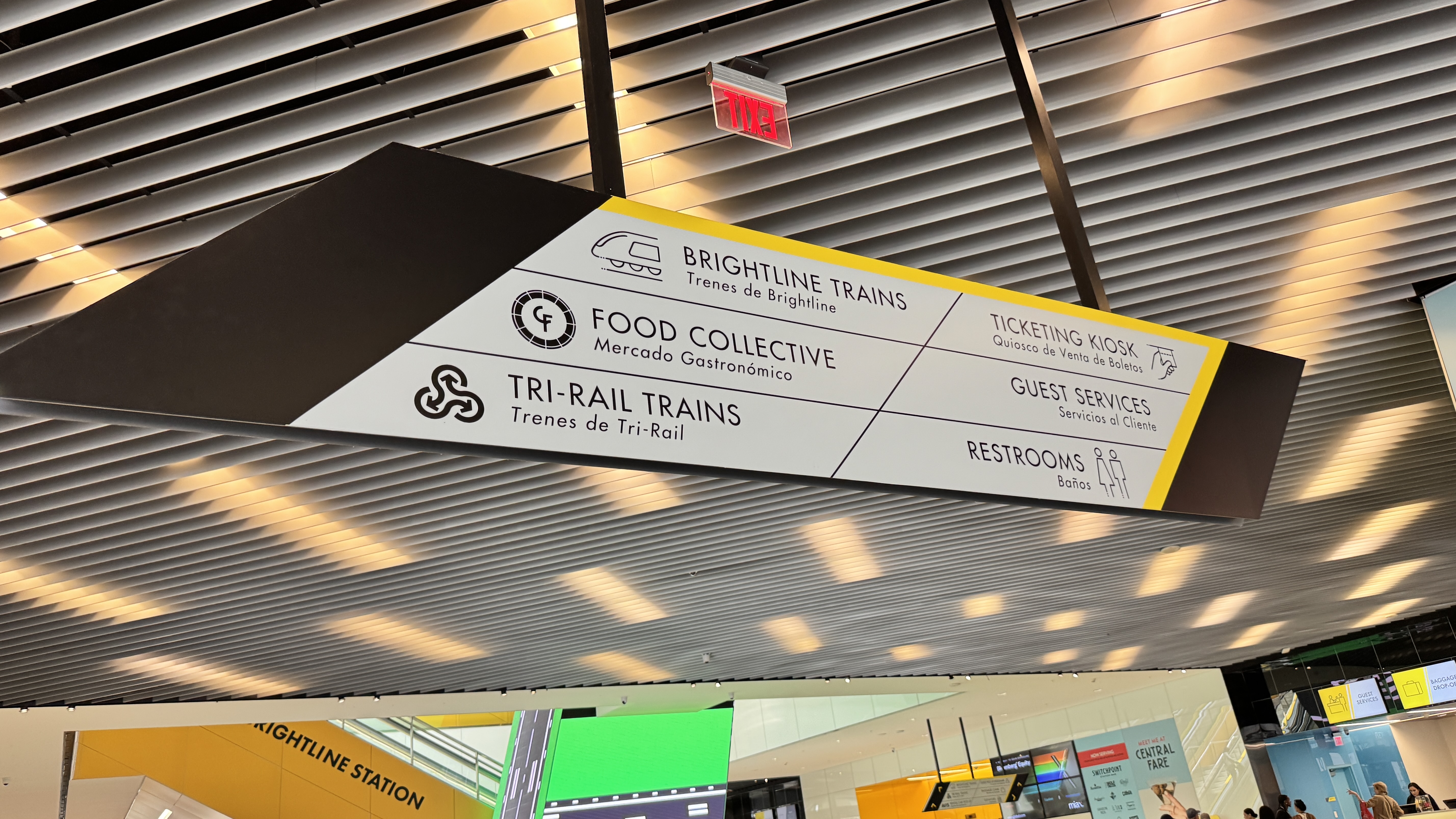 Placa de sinalização suspensa em um terminal, indicando direções para Brightline Trains, Food Collective, Tri-Rail Trains, Ticketing Kiosk, Guest Services e Restrooms, com texto em inglês e espanhol
