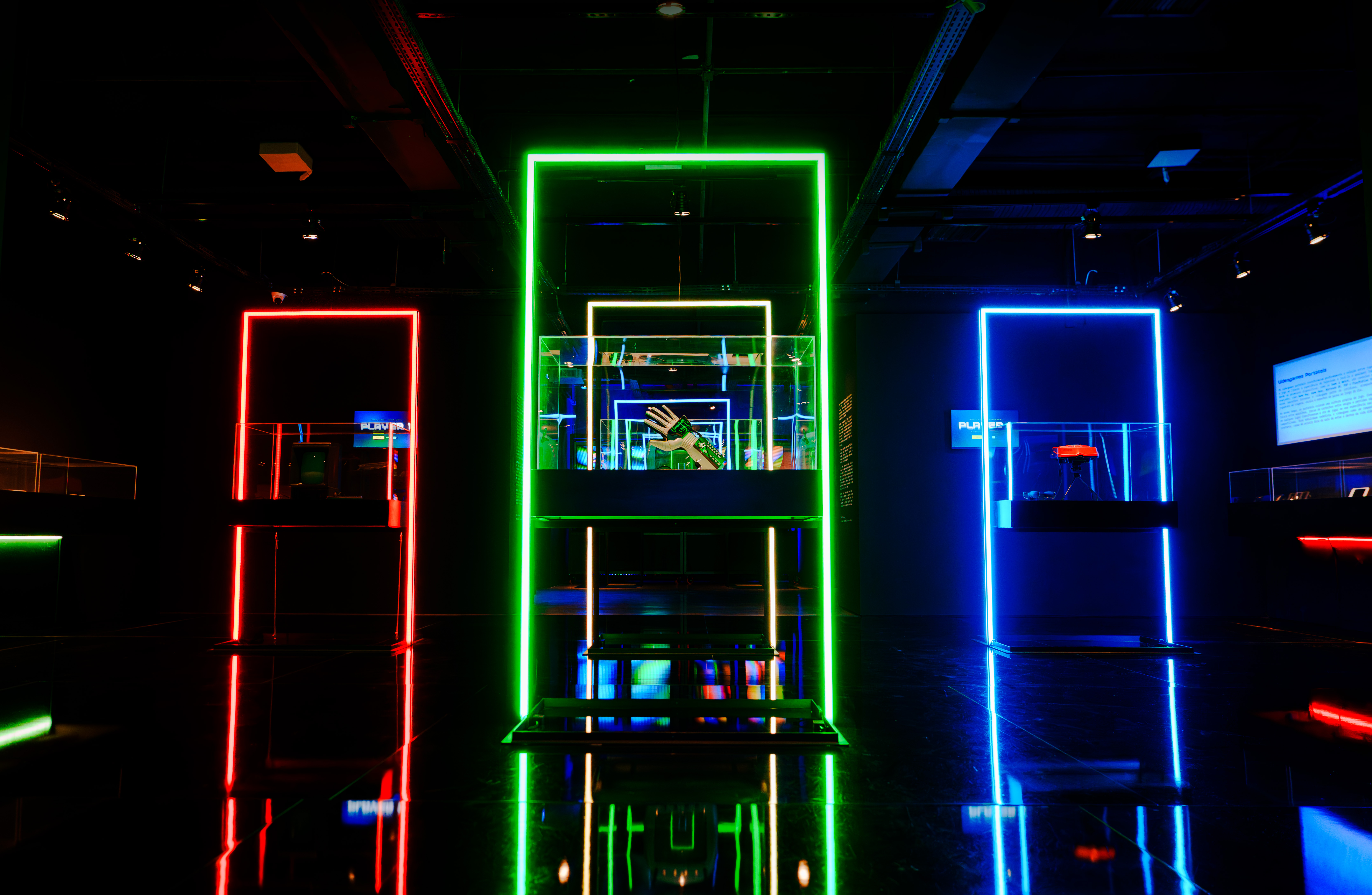 Exposição futurista com três estandes iluminados por neon: vermelho, verde e azul. O estande verde central exibe uma mão robótica dentro de uma caixa de vidro, refletindo no chão escuro.