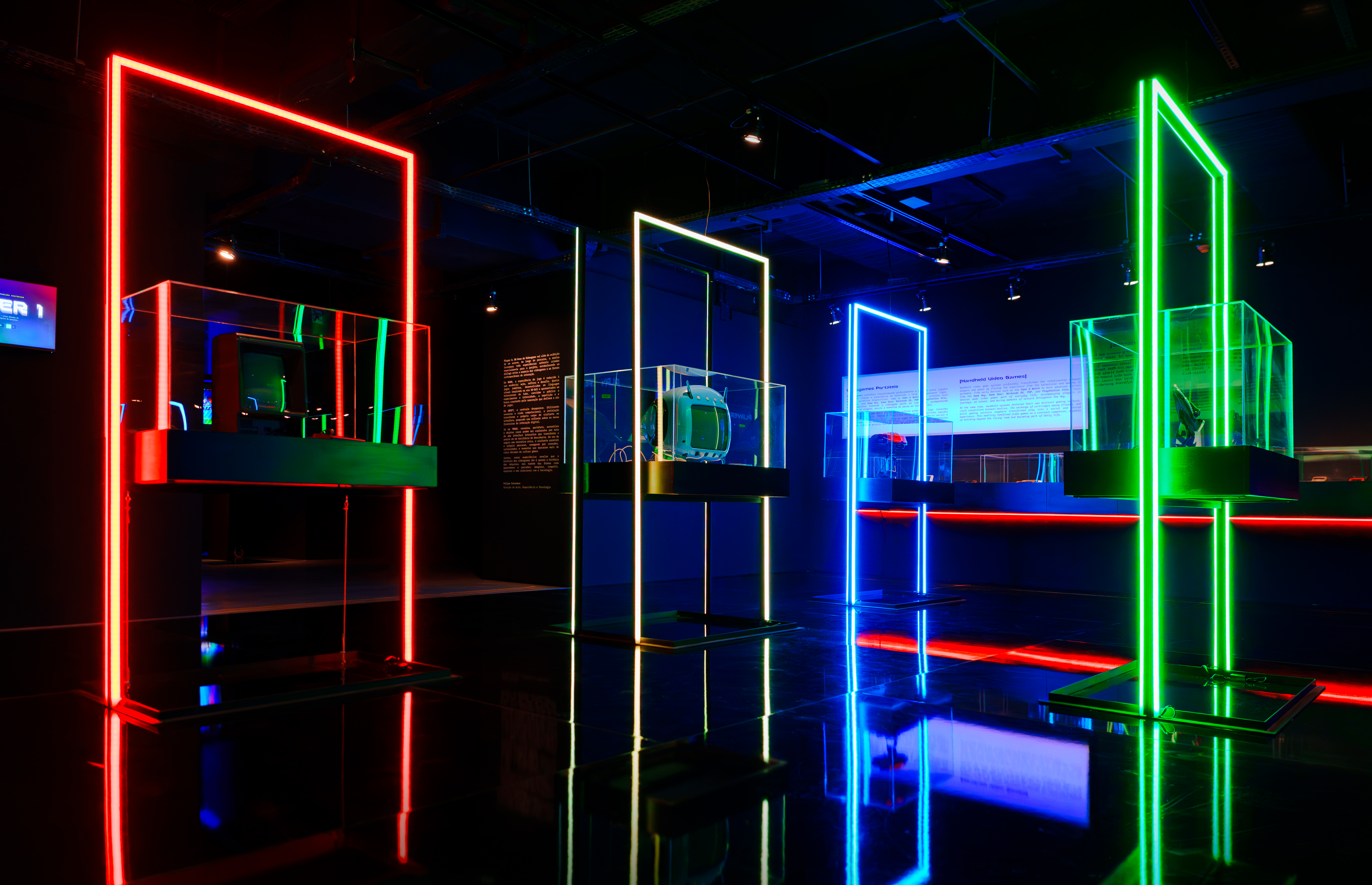 Exposição futurista com três displays iluminados por neon: vermelho à esquerda, amarelo no centro e verde à direita. Cada um exibe um objeto em uma caixa de vidro, com textos informativos ao fundo. O chão escuro reflete as luzes coloridas