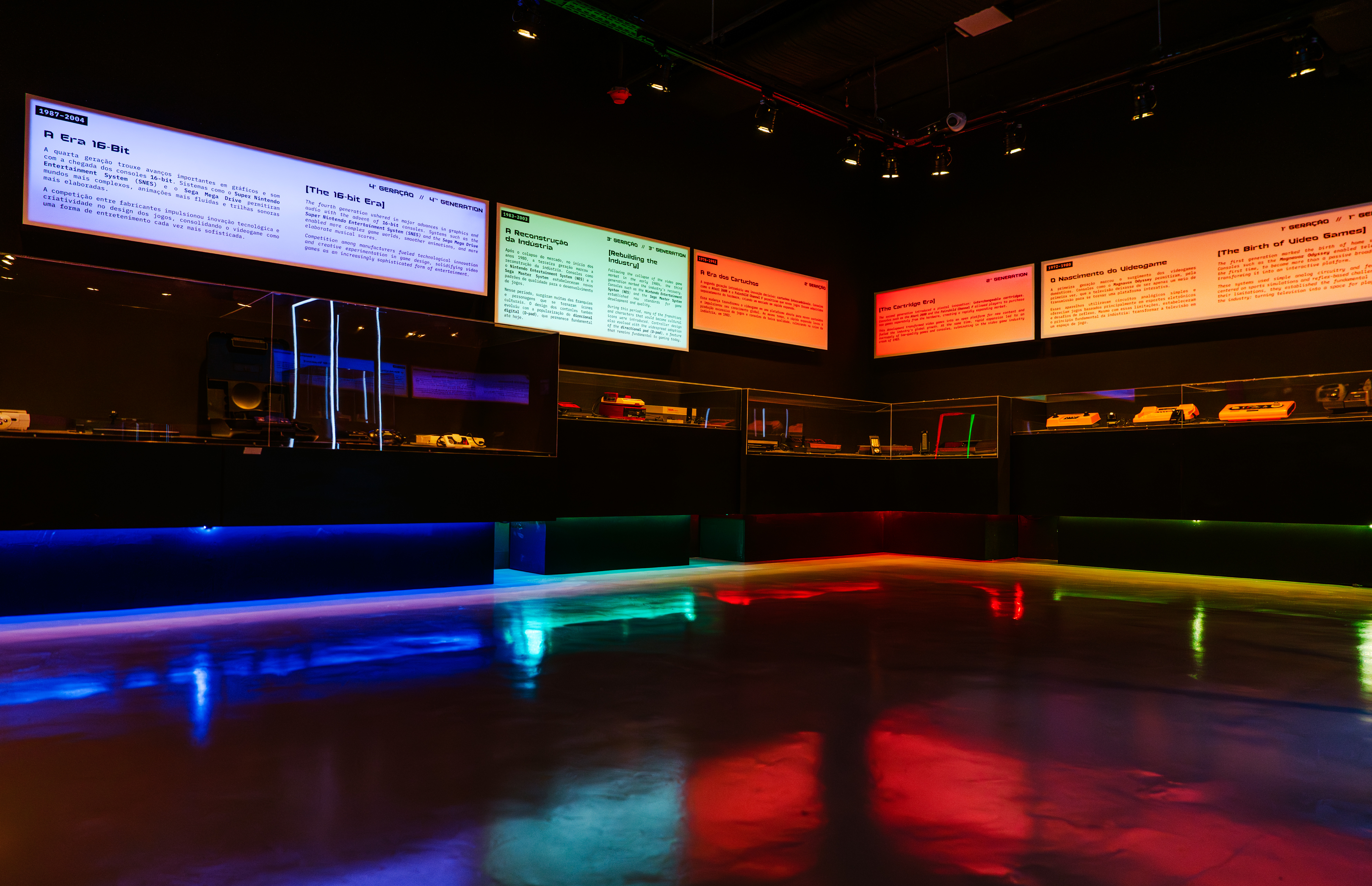 Exposição de videogames com painéis informativos iluminados em branco, verde e laranja, sobre a história dos jogos. Vitrines escuras exibem consoles e acessórios, enquanto o chão reflete luzes coloridas em tons de azul, verde e vermelho, criando um ambiente imersivo