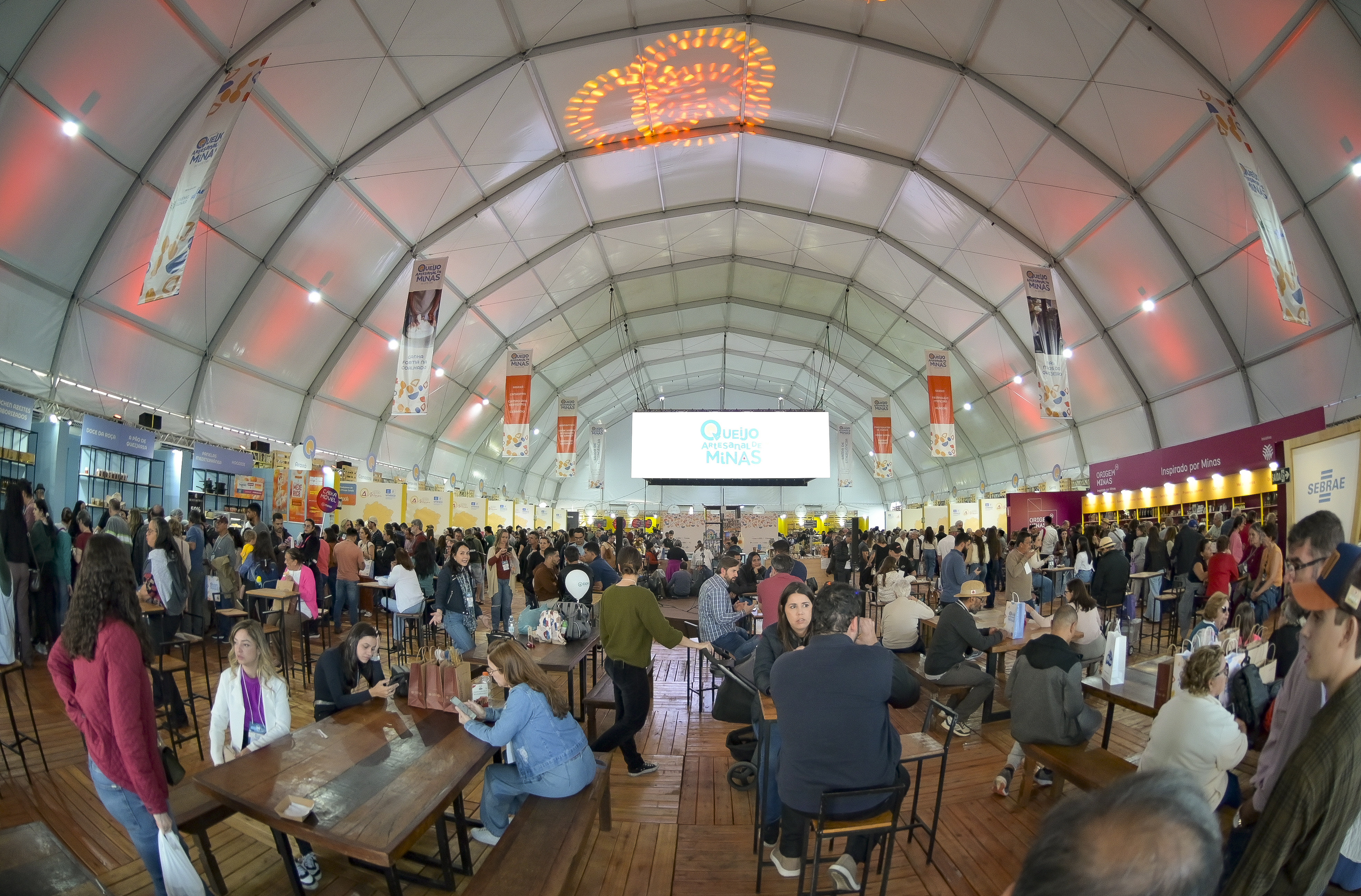 Grande tenda branca com centenas de pessoas em um evento, algumas sentadas em mesas de madeira, outras circulando. Um telão exibe 