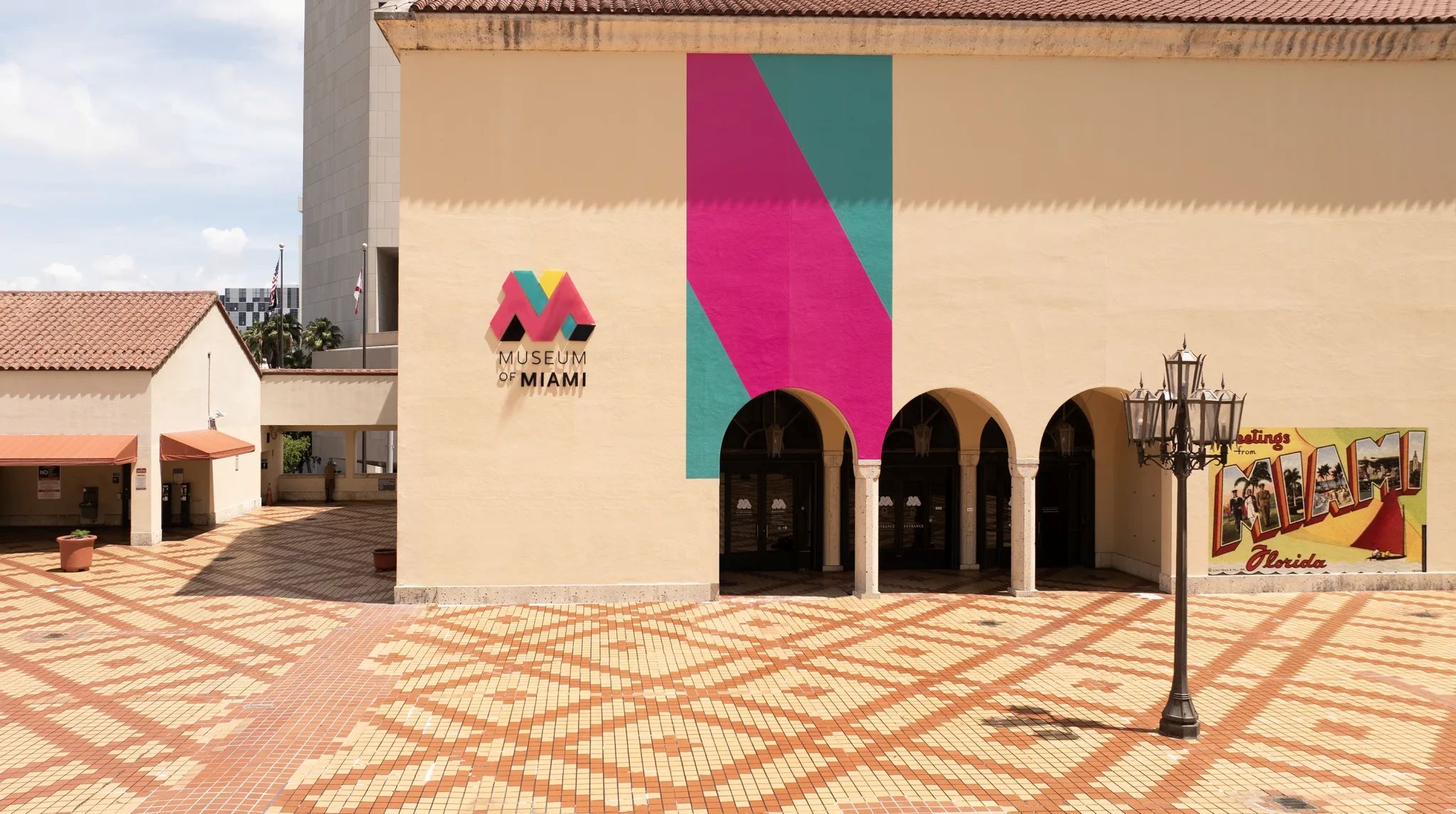 Fachada do Museum of Miami com logo colorido e um mural abstrato rosa e azul-petróleo. Três arcos escuros levam à entrada, e o pátio tem piso de tijolos em padrão geométrico. À direita, um poste de luz antigo e um mural estilo cartão-postal Greetings from Miami Florida.