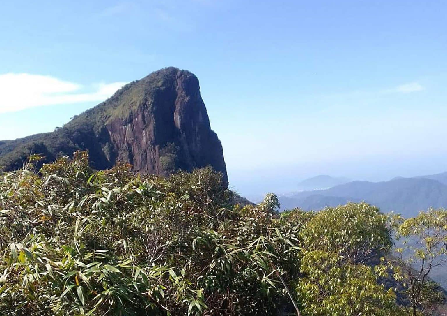 Montanha rochosa e íngreme, coberta por vegetação verde no topo, sob céu azul claro. Em primeiro plano, arbustos e folhagens densas. Ao fundo, outras montanhas e um vale com névoa.
