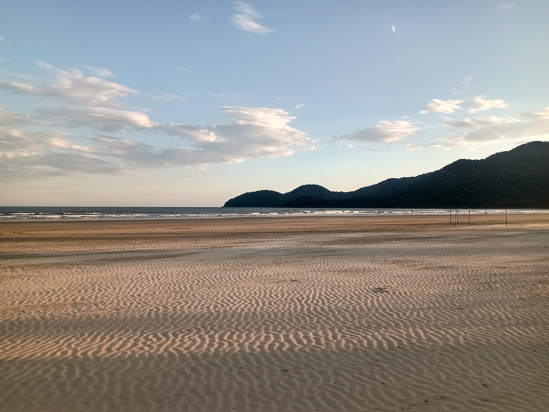 Praia deserta com areia clara e ondulada em primeiro plano, mar calmo ao centro e montanhas verdes escuras no horizonte sob um céu azul claro com nuvens esparsas
