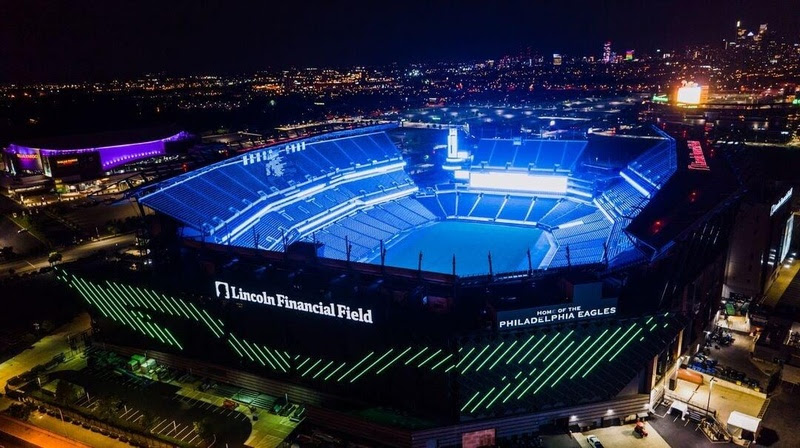 Vista aérea noturna do Lincoln Financial Field, estádio do Philadelphia Eagles, iluminado em azul vibrante. O campo e as arquibancadas brilham em azul, enquanto a fachada exibe o nome do estádio e HOME OF THE PHILADELPHIA EAGLES em branco, com linhas verdes diagonais. Ao fundo, a cidade se estende com luzes cintilantes