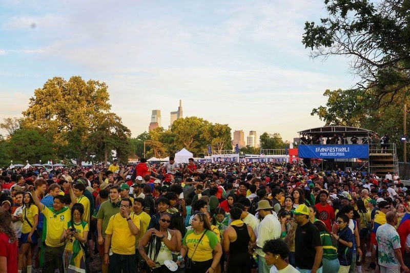 Multidão de pessoas em um festival ao ar livre, muitas vestindo camisas amarelas e verdes, com um palco e edifícios ao fundo