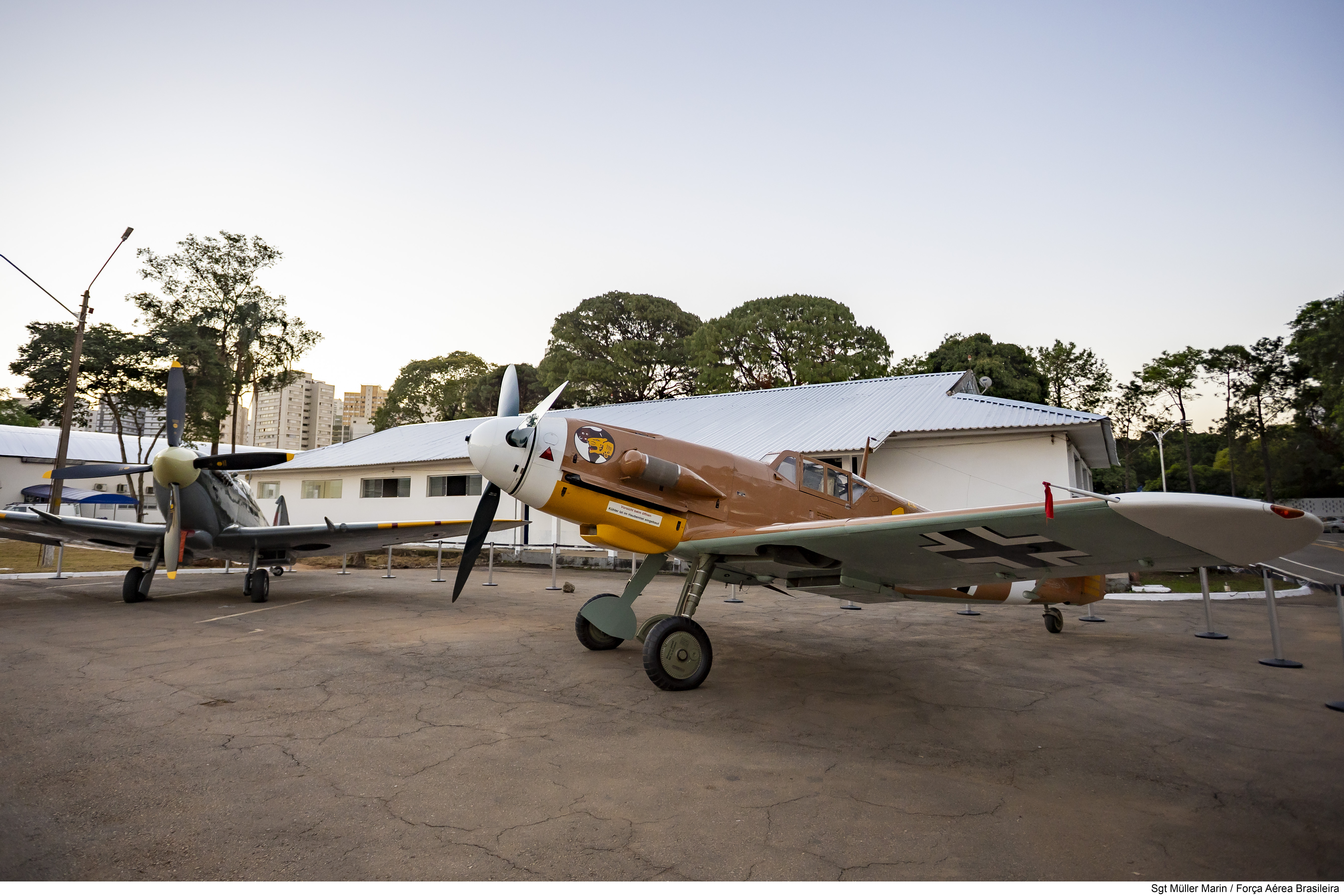 Dois aviões de guerra antigos em exposição, um cinza à esquerda e um marrom-amarelado à direita, com hélices e asas marcadas com cruzes pretas, em um pátio de concreto com prédios brancos e árvores ao fundo