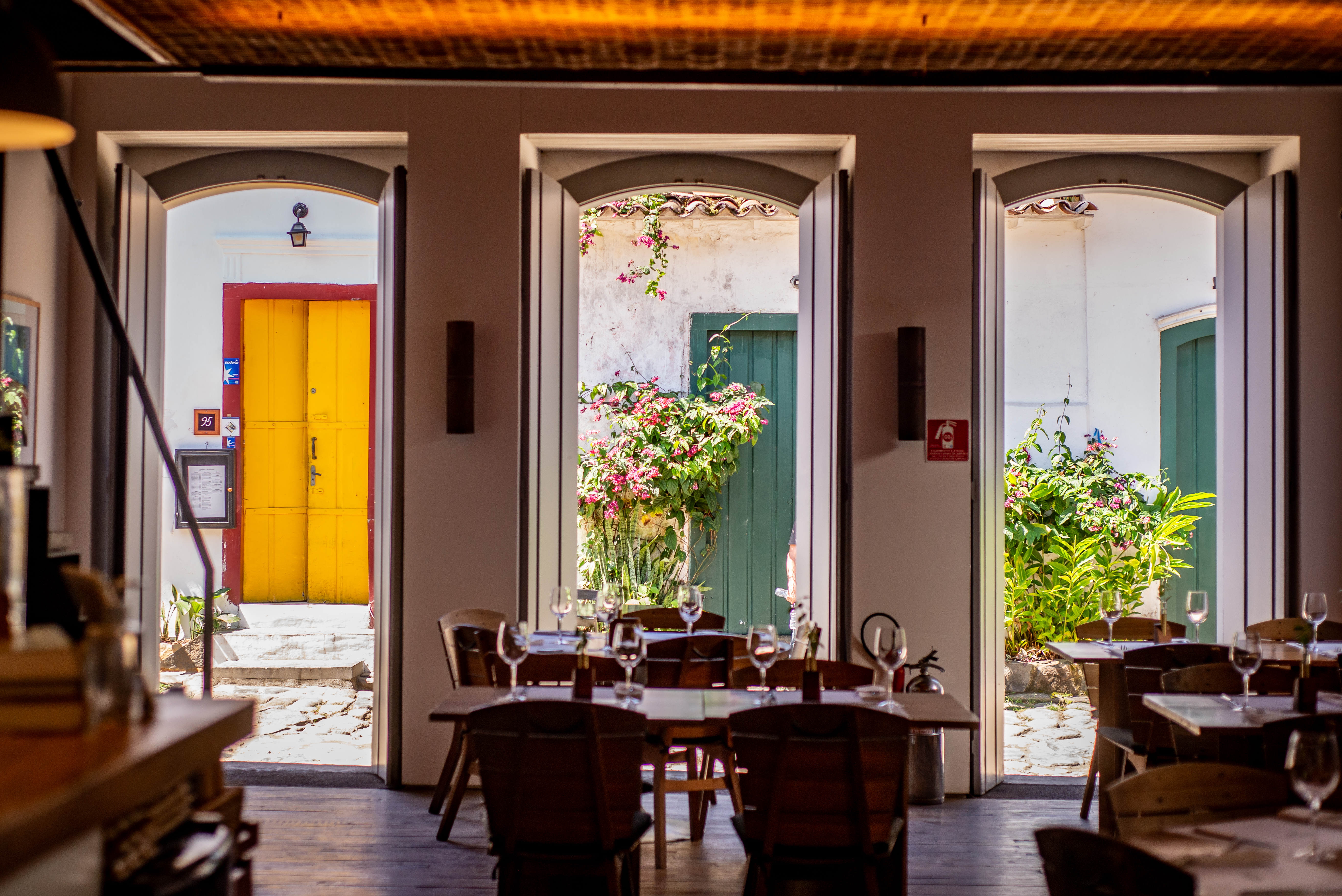 Interior de um restaurante com mesas de madeira e cadeiras escuras, com taças de vinho. Três portas arqueadas revelam um pátio externo com plantas e flores, e uma porta amarela vibrante ao fundo. O chão é de madeira escura e o teto tem um acabamento rústico