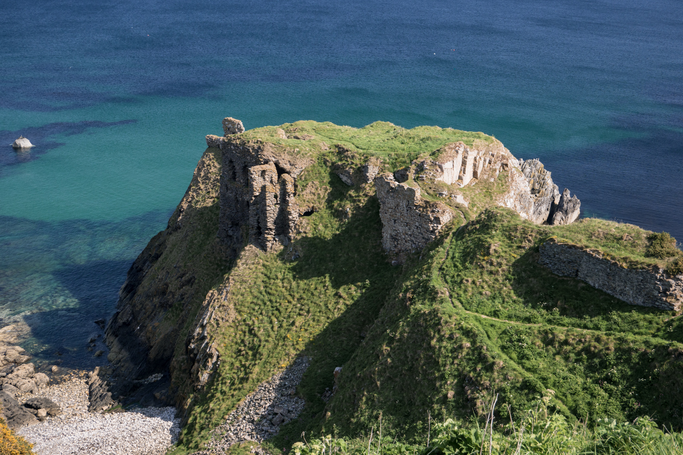 Ruínas de um castelo antigo no topo de um penhasco rochoso coberto por grama verde, com o mar azul-turquesa e azul-escuro ao fundo e uma praia de pedras na base