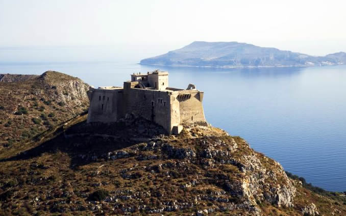 Antigo forte de pedra em um morro rochoso com vegetação rasteira, com o mar azul e uma ilha montanhosa ao fundo sob céu claro