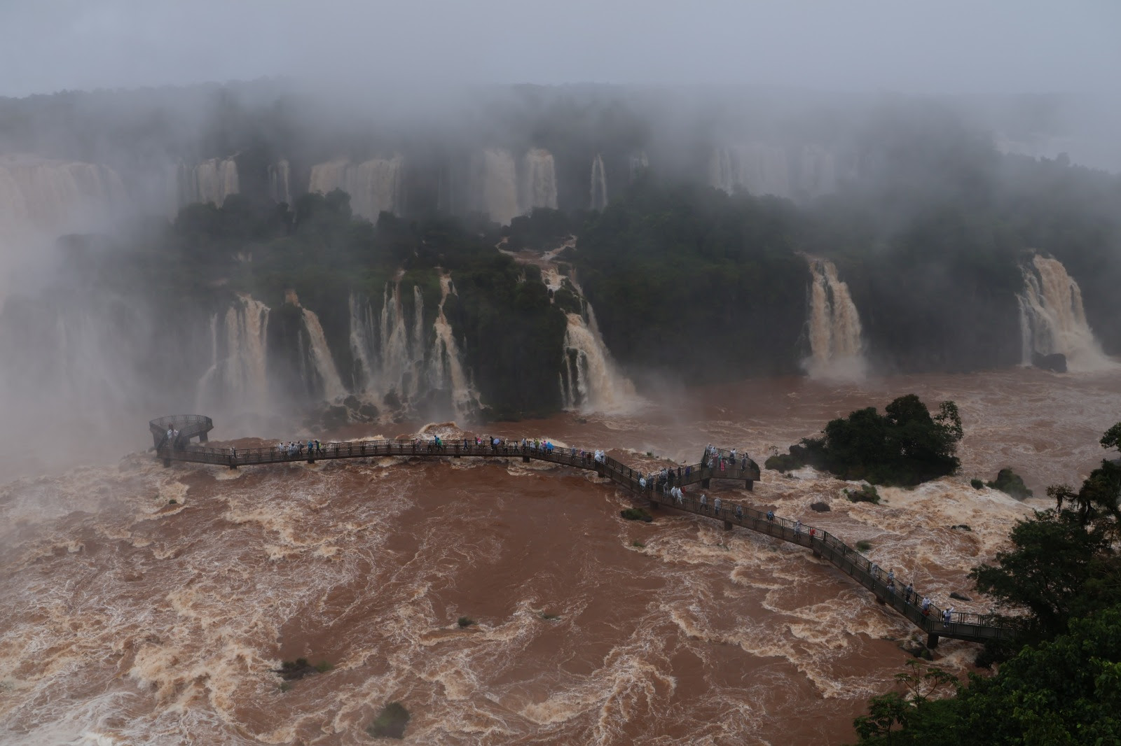 Vista aérea das Cataratas do Iguaçu em dia nublado, com névoa cobrindo o topo das quedas. A água barrenta do rio corre forte, e uma passarela de madeira com muitos turistas se estende sobre o rio, levando a uma plataforma de observação próxima às quedas. A vegetação verde densa emoldura a paisagem