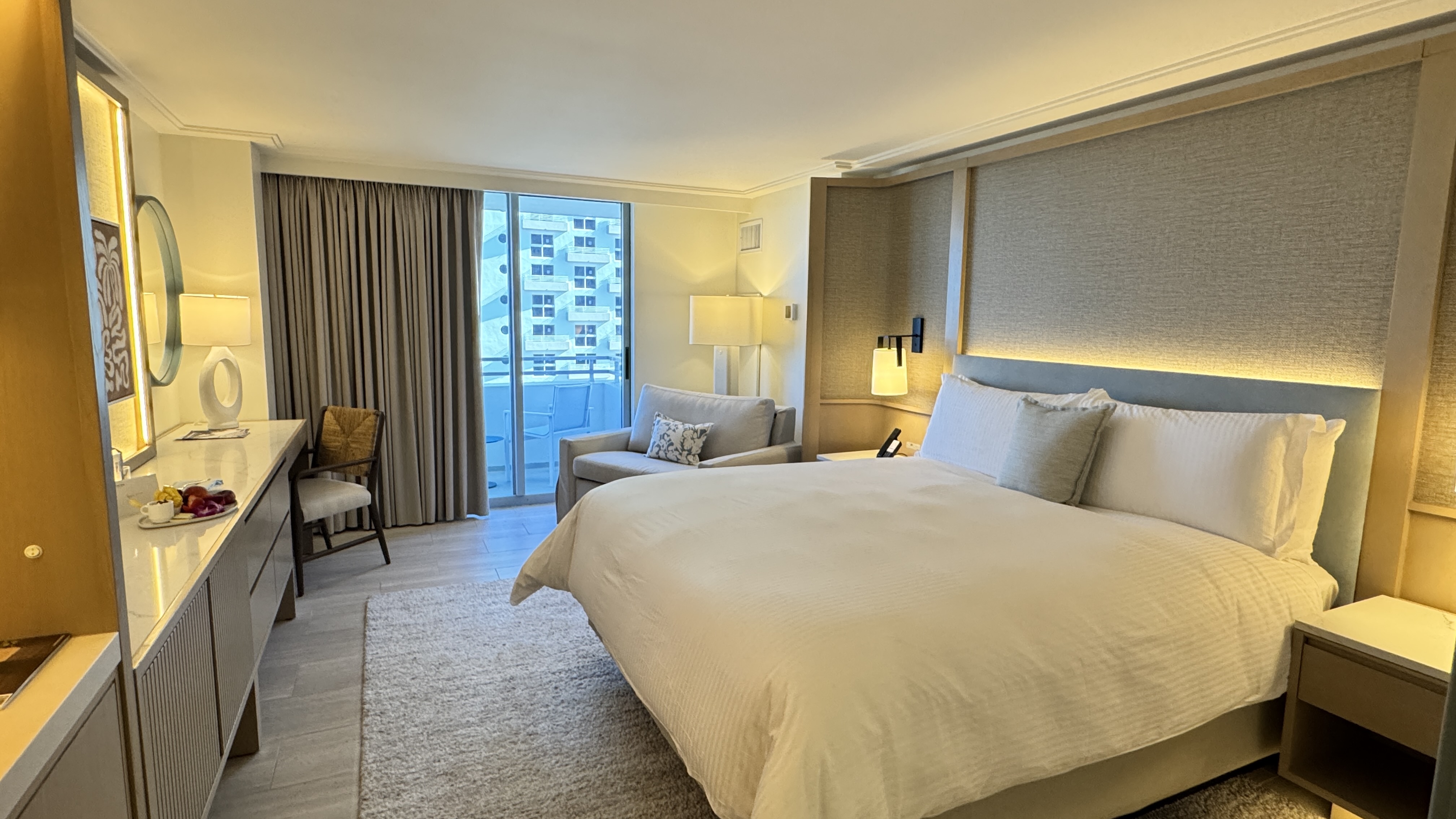 Quarto de hotel luxuoso com cama king-size de lençóis brancos, cabeceira estofada e iluminação indireta. À direita, uma mesa de cabeceira com luminária. À esquerda, um sofá cinza, uma poltrona de madeira e uma bancada com espelho, fruteira e luminária. Ao fundo, uma porta de vidro dá para uma varanda com vista para prédios. O ambiente tem piso claro e carpete felpudo.