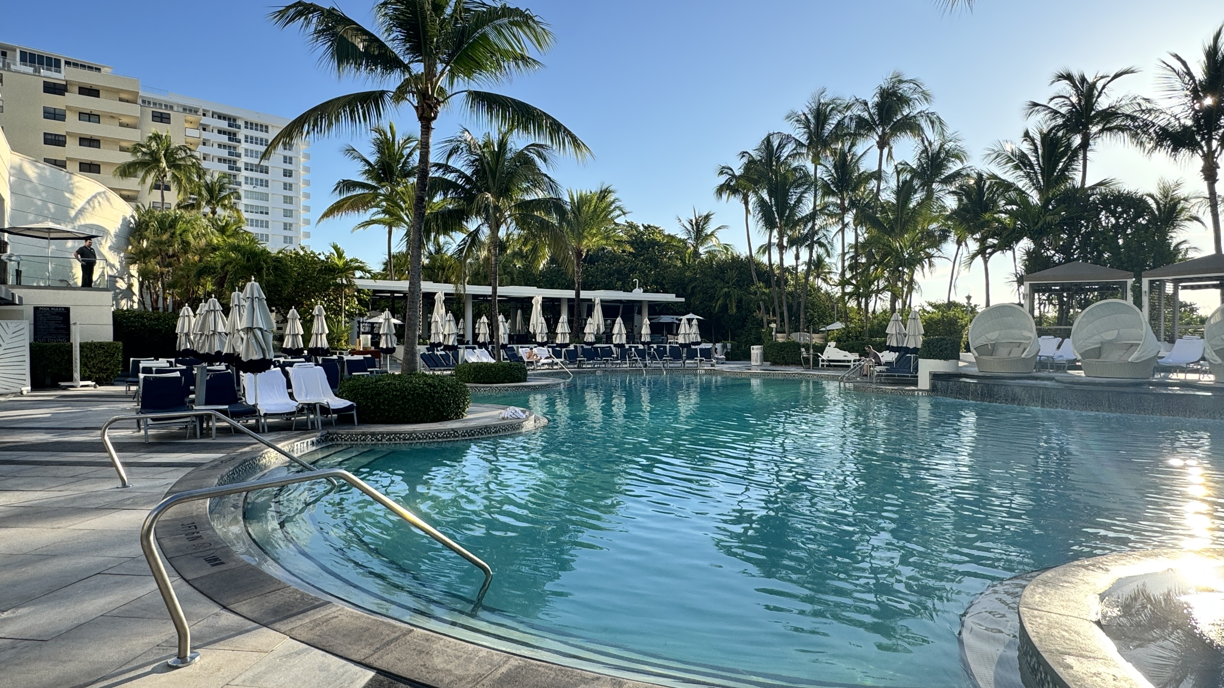 Piscina de hotel com água azul-turquesa, rodeada por espreguiçadeiras, guarda-sóis brancos e palmeiras. Ao fundo, prédios altos e o sol refletindo na água, criando um ambiente de lazer e relaxamento. Um homem observa da varanda à esquerda