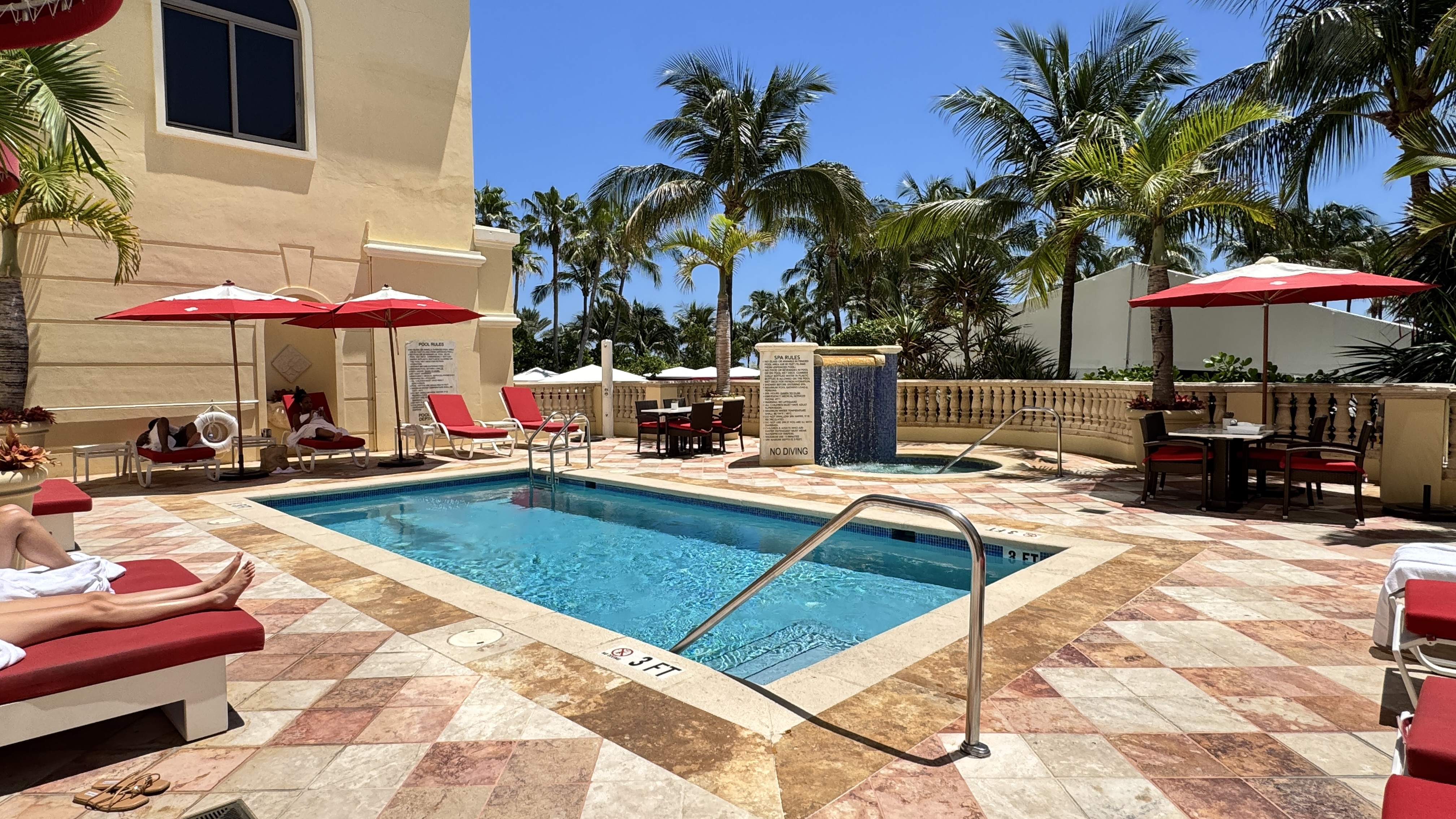 Piscina retangular de hotel com água azul, cercada por espreguiçadeiras vermelhas e guarda-sóis. Palmeiras altas e um prédio bege com janelas escuras ao fundo, sob céu azul claro. Uma pessoa relaxa em uma espreguiçadeira à esquerda.