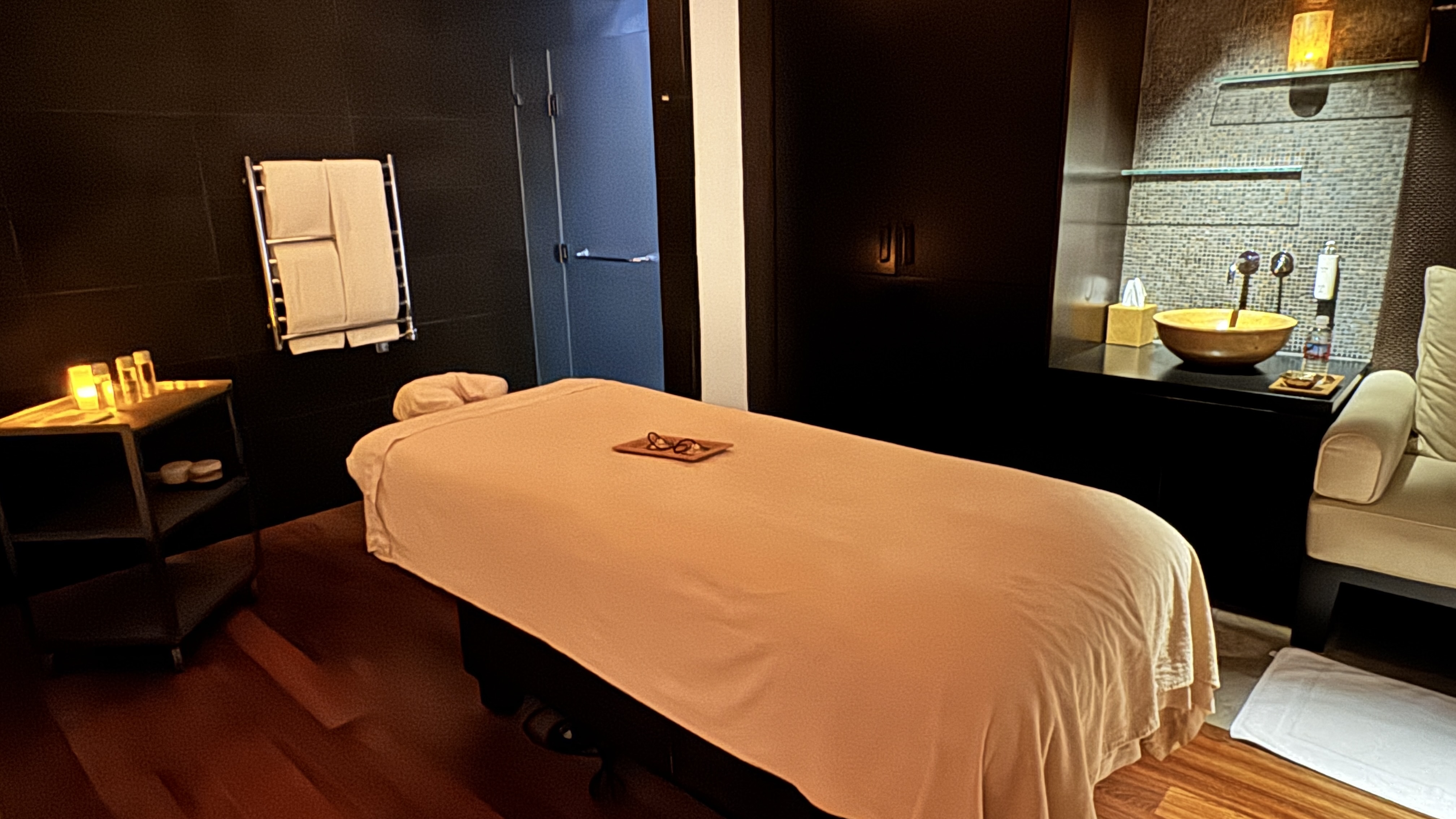 Sala de spa com iluminação suave, maca de massagem coberta por lençol pêssego, óculos sobre bandeja de madeira. À esquerda, mesa de apoio com velas acesas e toalhas em aquecedor. À direita, pia dourada sobre balcão escuro e poltrona creme. O ambiente é relaxante e acolhedor