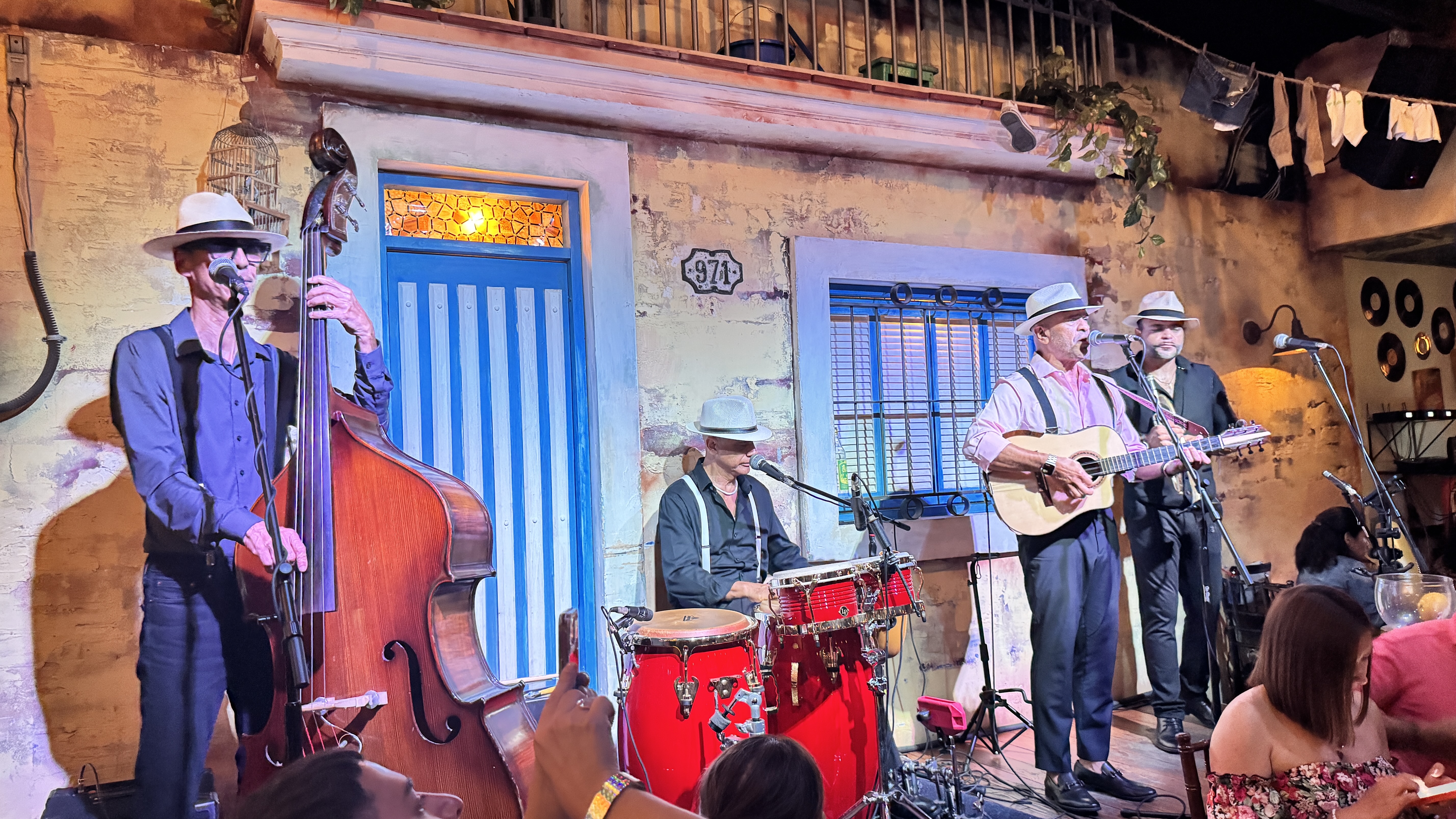 Quatro músicos homens, todos de chapéu, tocam instrumentos em um palco decorado como uma fachada de casa antiga. Um toca contrabaixo, outro bateria, e dois violão, cantando em microfones. O público assiste em primeiro plano