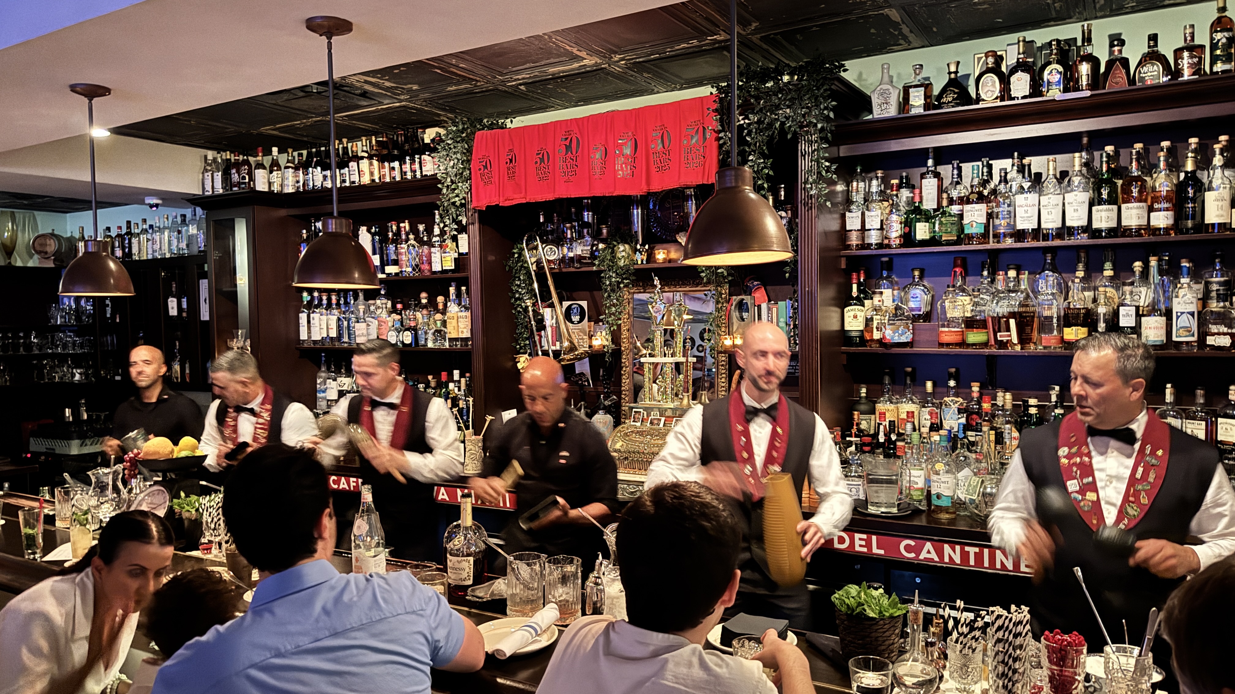 Seis bartenders, cinco homens e uma mulher, preparam bebidas em um bar com prateleiras cheias de garrafas de bebidas alcoólicas. Dois homens usam coletes pretos com laços vermelhos e medalhas, enquanto os outros usam camisas pretas. Clientes sentam-se à frente do balcão, observando a preparação. O ambiente é iluminado por pendentes de teto e decorado com folhagens e um espelho dourado. Uma faixa vermelha com caracteres dourados decora a parede.