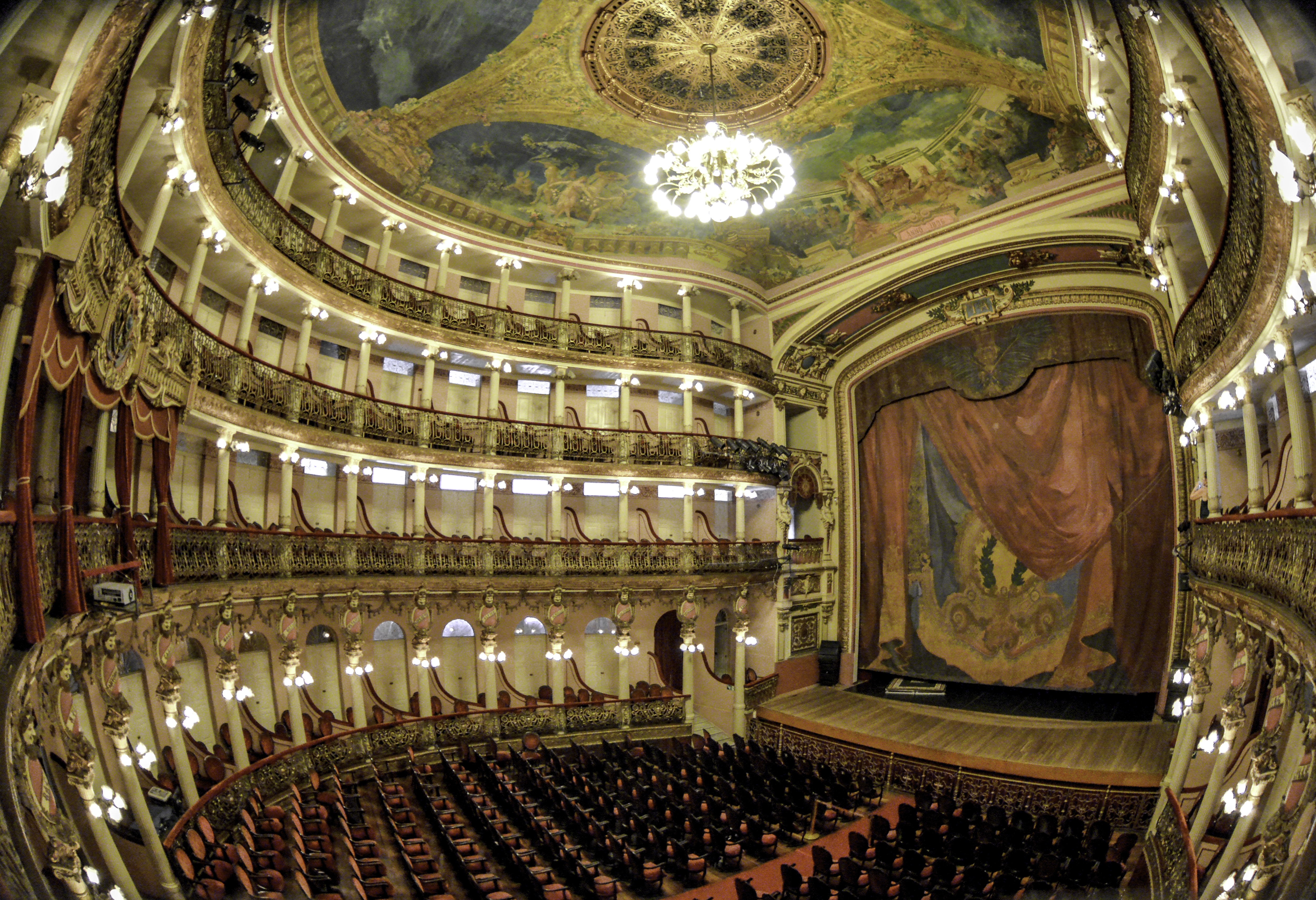 Interior de um teatro opulento com poltronas vermelhas e cinco andares de camarotes dourados e rosados. Um lustre central ilumina o teto pintado com cenas mitológicas. O palco tem uma cortina vermelha e azul com detalhes dourados. A imagem é capturada com uma lente olho de peixe, distorcendo as laterais.