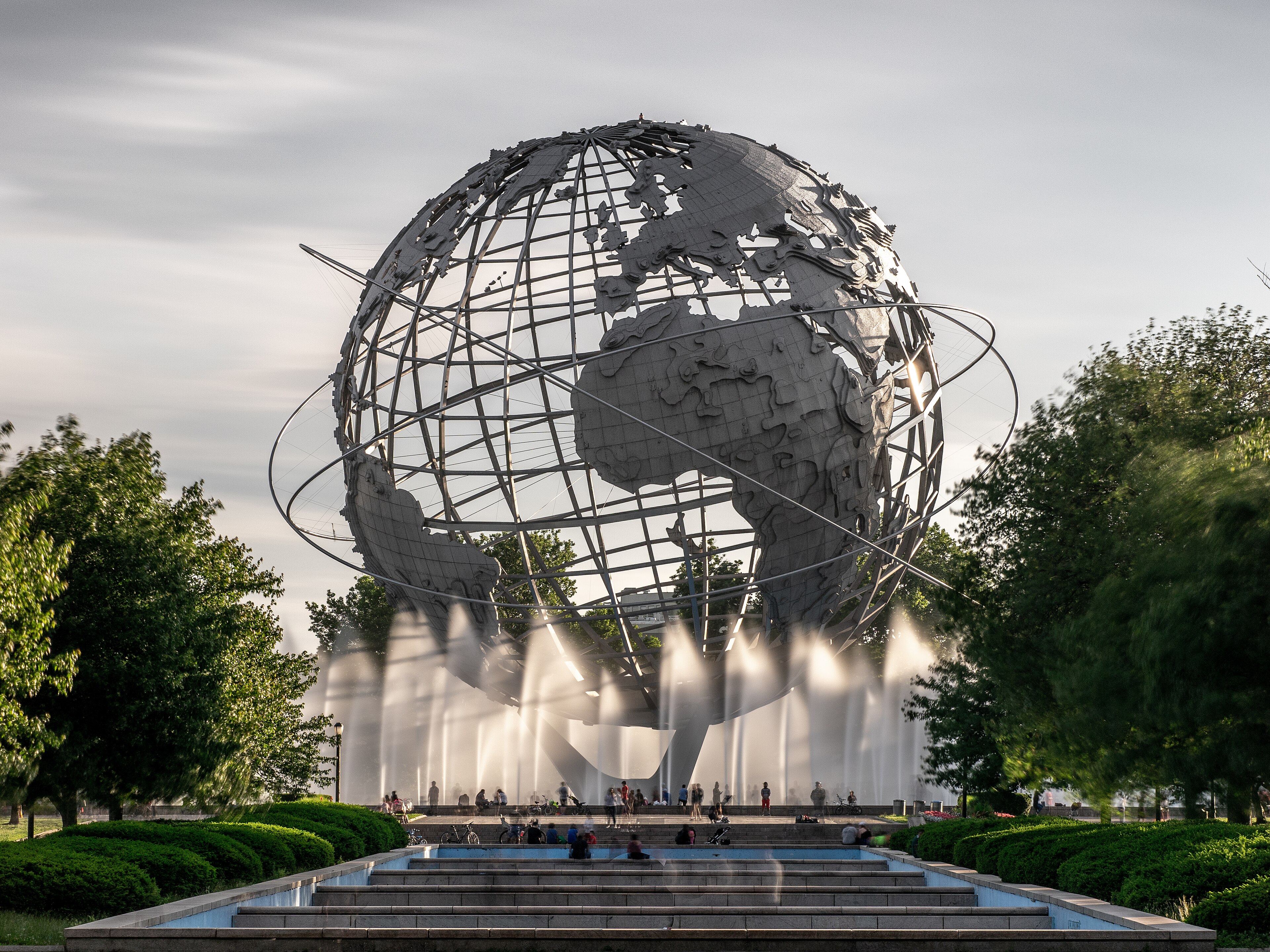 A Unisphere, um globo terrestre gigante de metal, com jatos dágua e pessoas ao redor, em um parque com árvores e arbustos verdes sob céu claro