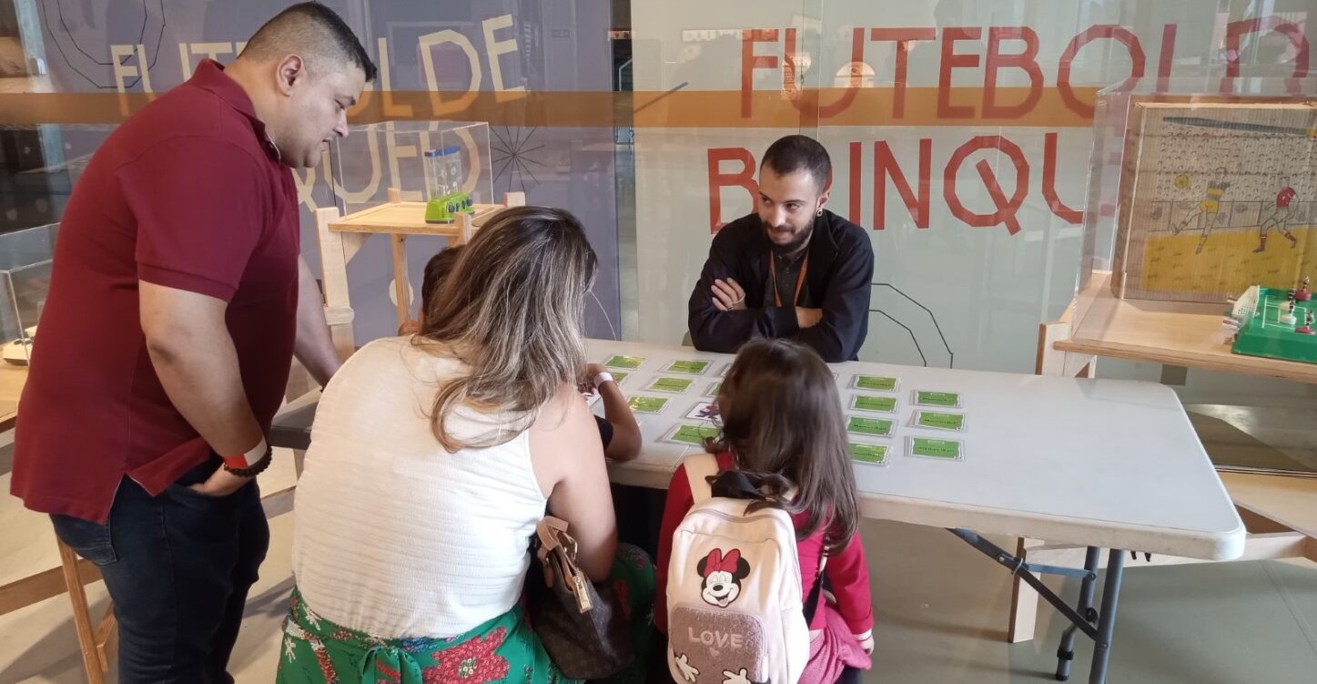 Três adultos e uma criança interagem em uma mesa com cartas verdes. Um homem de camisa polo vermelha e outro de jaqueta preta observam, enquanto uma mulher de blusa branca e uma menina de mochila da Minnie com a palavra LOVE olham para as cartas. Ao fundo, uma vitrine exibe um jogo de futebol de botão e um painel com a palavra FUTEBOL