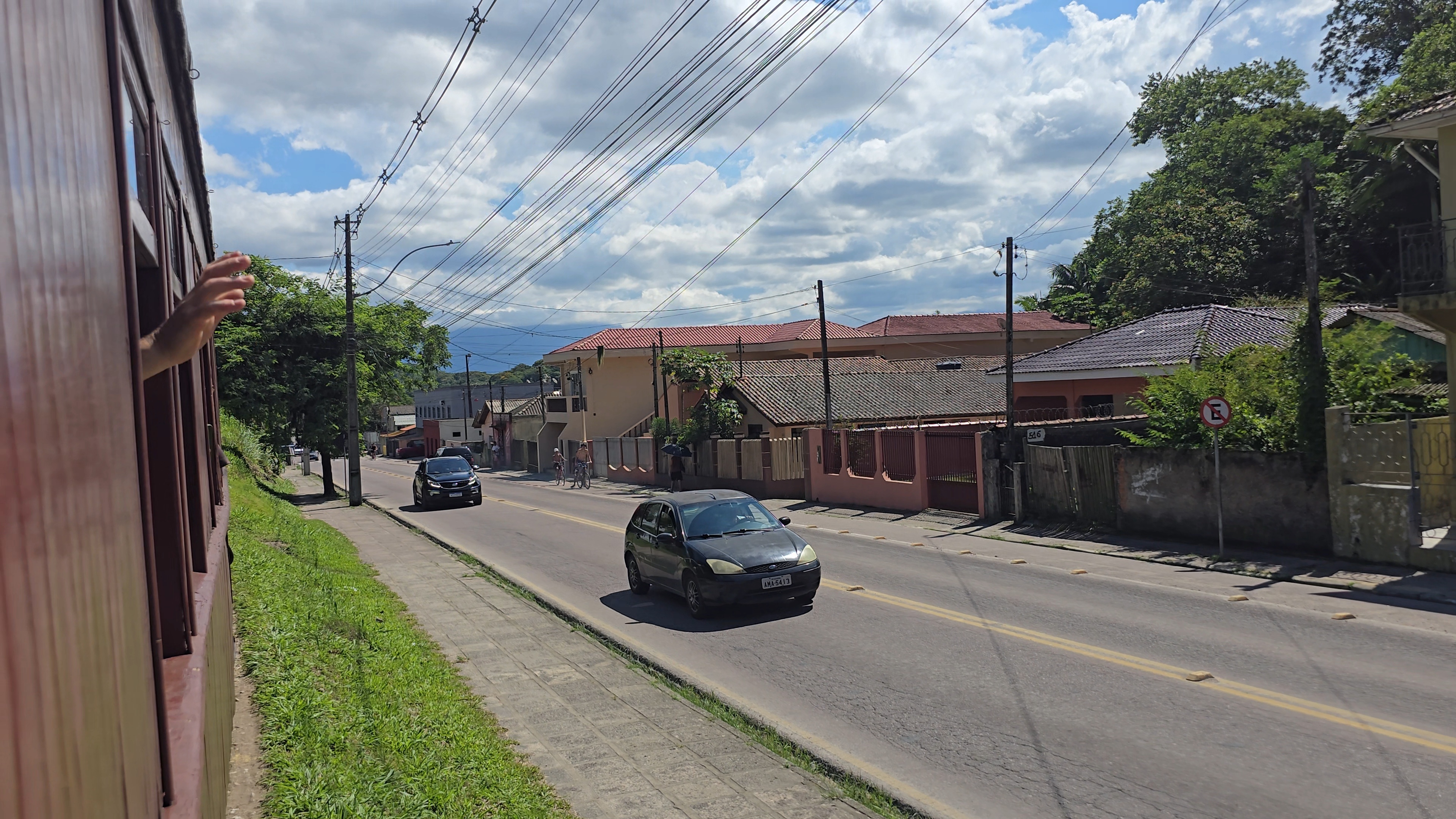 Mão de uma pessoa acenando pela janela de um trem em movimento, com uma rua paralela à direita, carros, casas e vegetação sob céu azul com nuvens