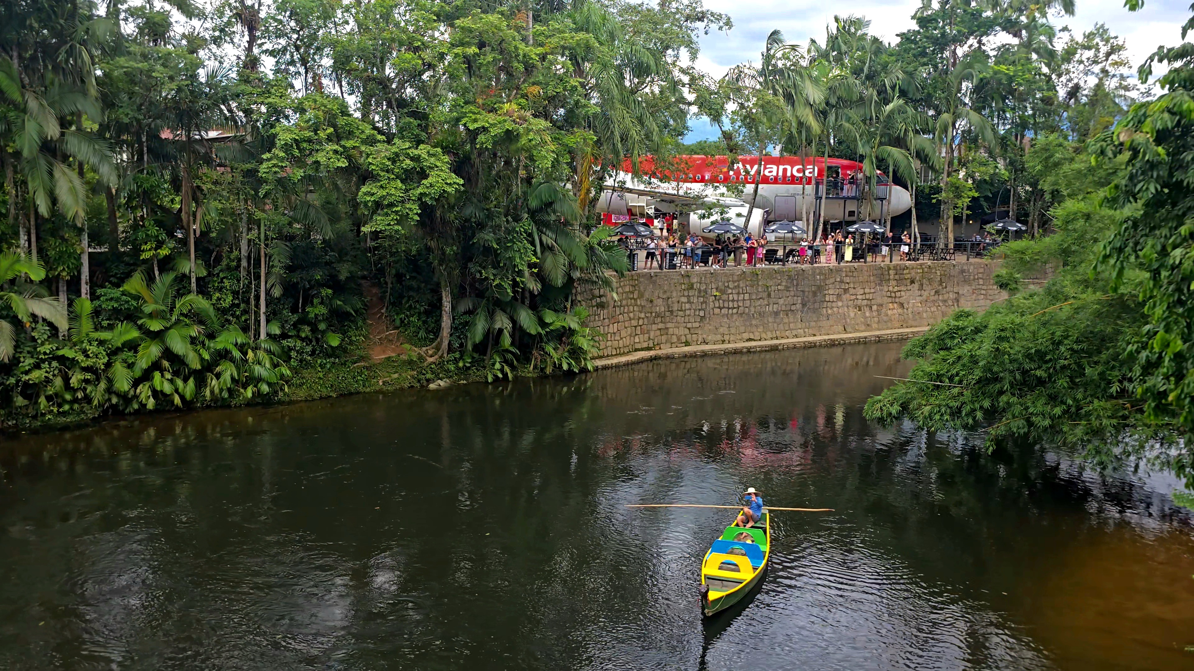 Um homem em um chapéu de palha rema um barco colorido (amarelo, azul e verde) em um rio escuro. À direita, um muro de pedra sustenta uma área com pessoas e a fuselagem de um avião vermelho e branco da Avianca, cercado por densa vegetação tropical