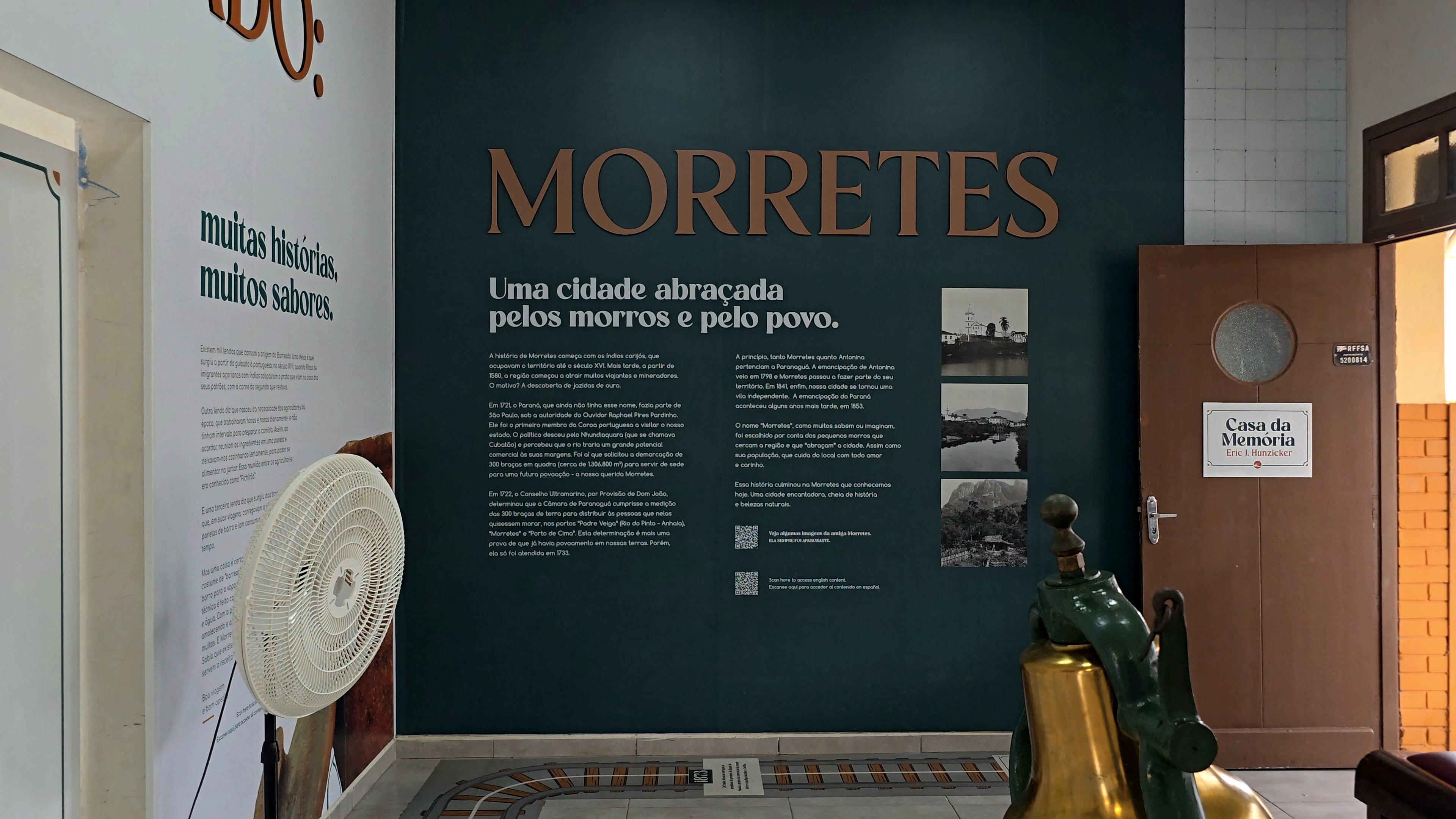 Parede verde-escura com texto MORRETES Uma cidade abraçada pelos morros e pelo povo e três fotos em preto e branco de paisagens. À esquerda, parede branca com texto muitas histórias, muitos sabores e um ventilador branco. À direita, porta marrom com placa Casa da Memória. No chão, trilhos de trem em miniatura e um sino dourado e verde