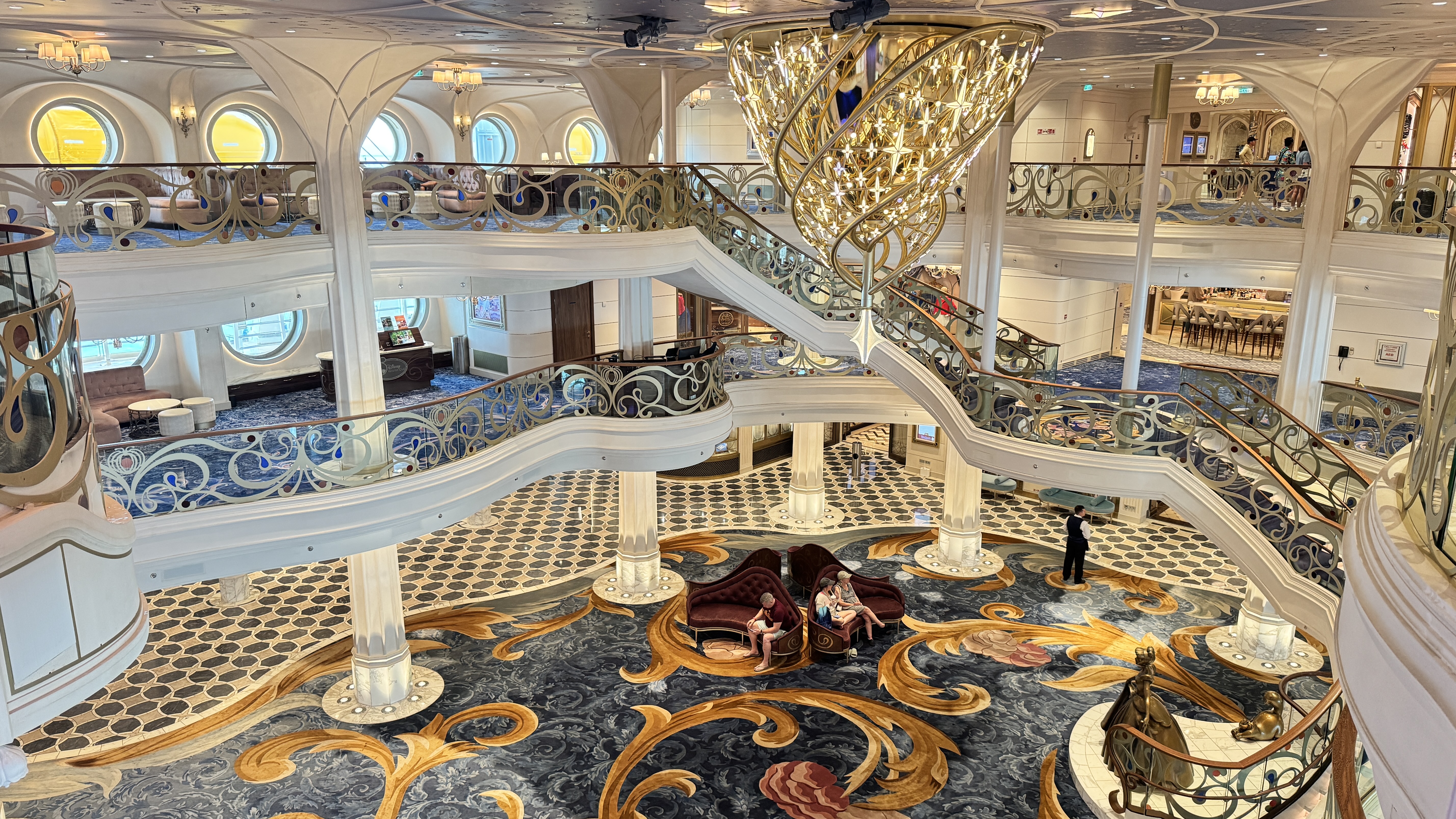 Interior luxuoso de um navio, com escadarias brancas e corrimãos dourados ornamentados. Um lustre dourado imponente pende do teto. No térreo, um tapete azul escuro com detalhes dourados e vermelhos, onde pessoas sentam em sofás curvos de veludo vermelho. Outras pessoas são vistas nos andares superiores, em áreas de convivência com janelas redondas e sofás. Um homem de terno preto está em pé no térreo, próximo a uma estátua dourada. O ambiente é grandioso e elegante