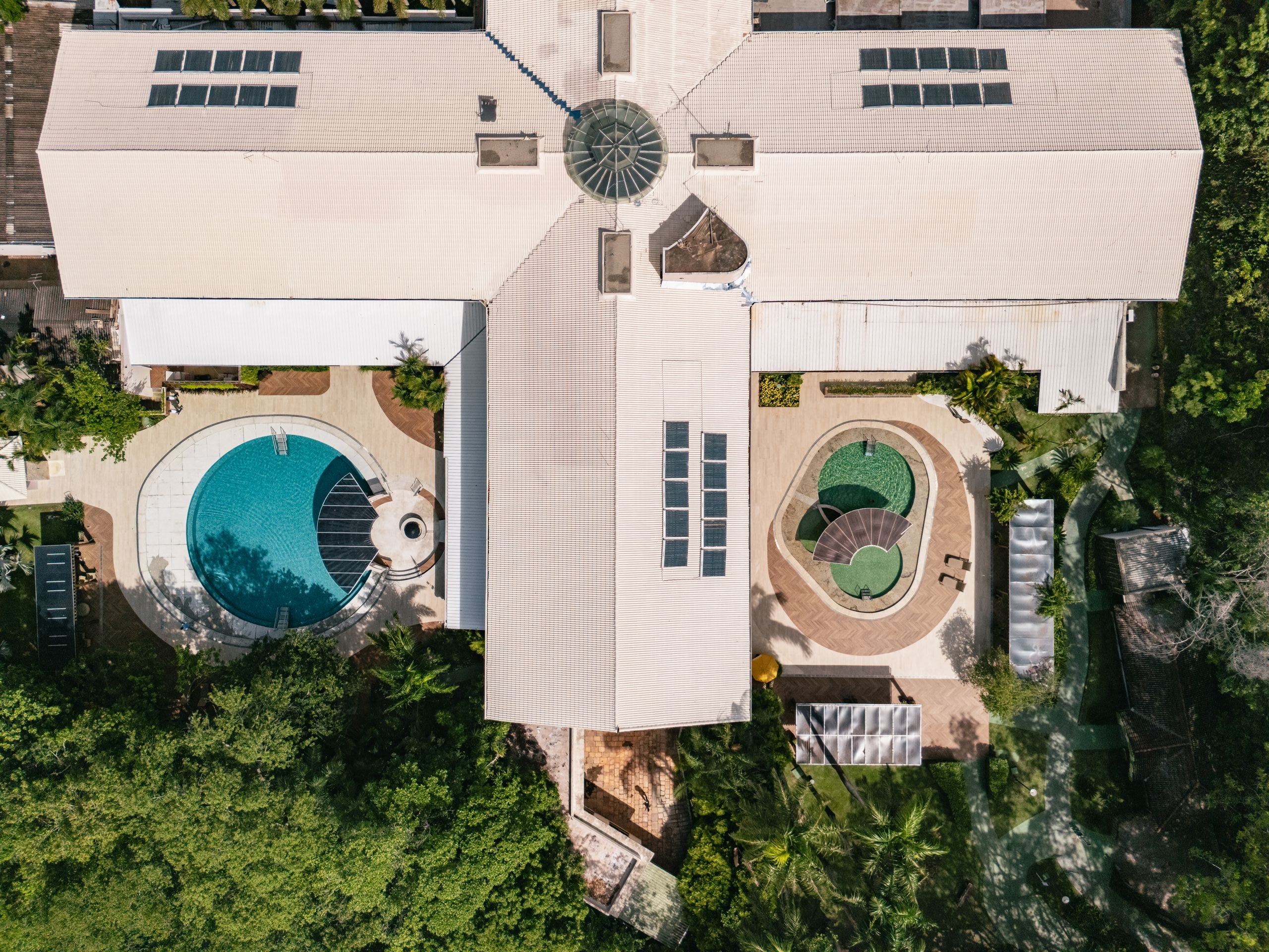 Vista aérea de um complexo de lazer com telhados claros, duas piscinas e muita vegetação. À esquerda, uma piscina azul em formato de C com deck de madeira. À direita, duas piscinas menores de formato oval com água esverdeada. Painéis solares e claraboias no telhado complementam a estrutura.