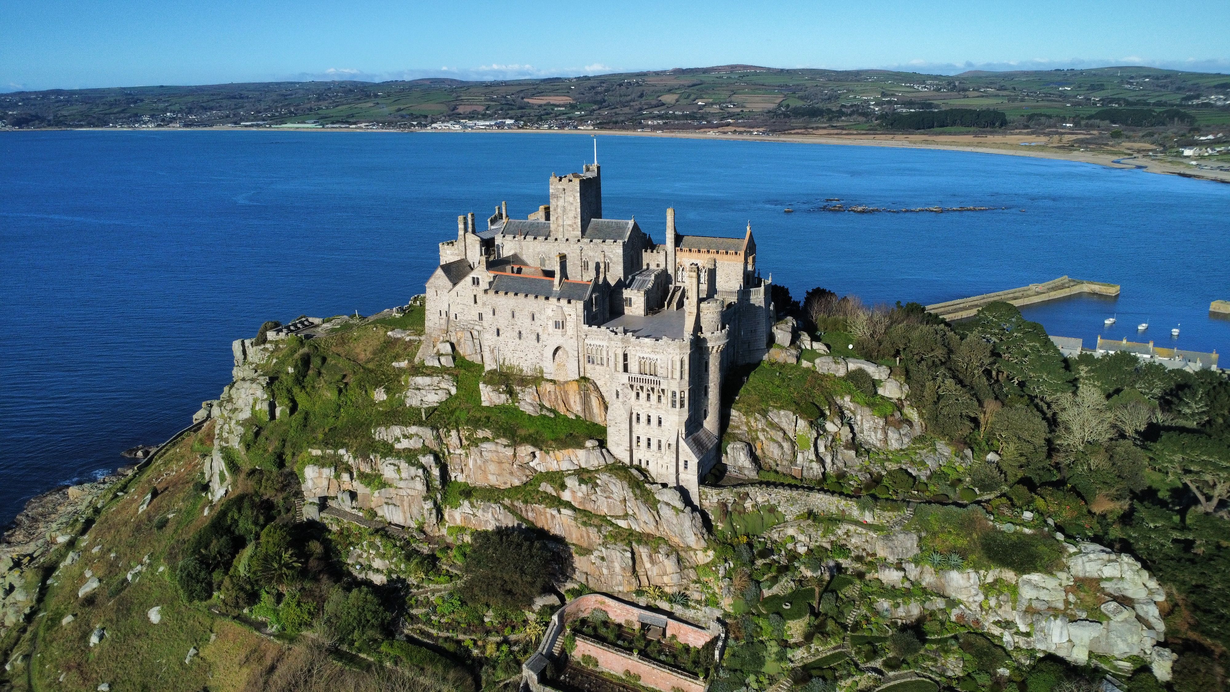 Castelo histórico de pedra cinza com torres e telhados escuros, construído sobre uma ilha rochosa e verdejante. Ao fundo, um mar azul intenso e uma faixa de terra com vegetação e casas. À direita, um pequeno porto com barcos brancos.