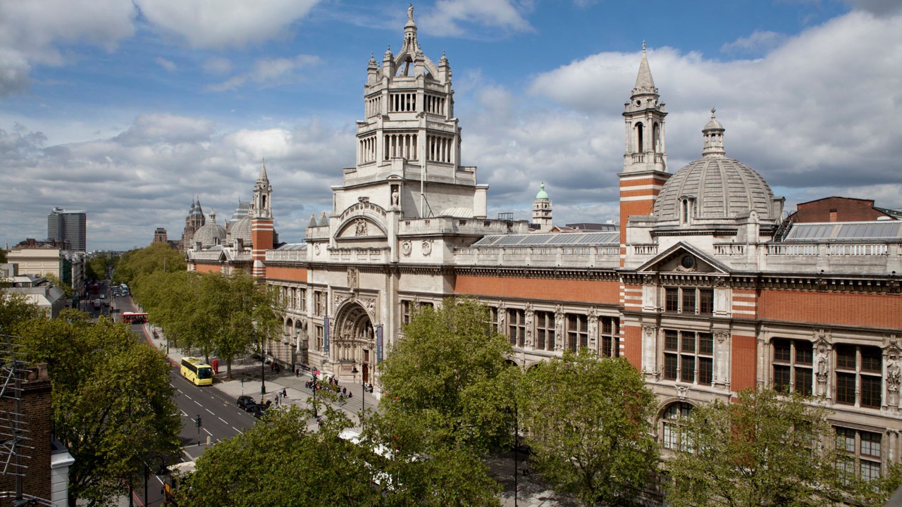 Vista aérea do Victoria and Albert Museum em Londres, com sua fachada de tijolos vermelhos e detalhes em pedra clara, incluindo uma torre central alta e uma cúpula. Uma rua arborizada com ônibus e carros passa em frente ao museu, sob um céu parcialmente nublado