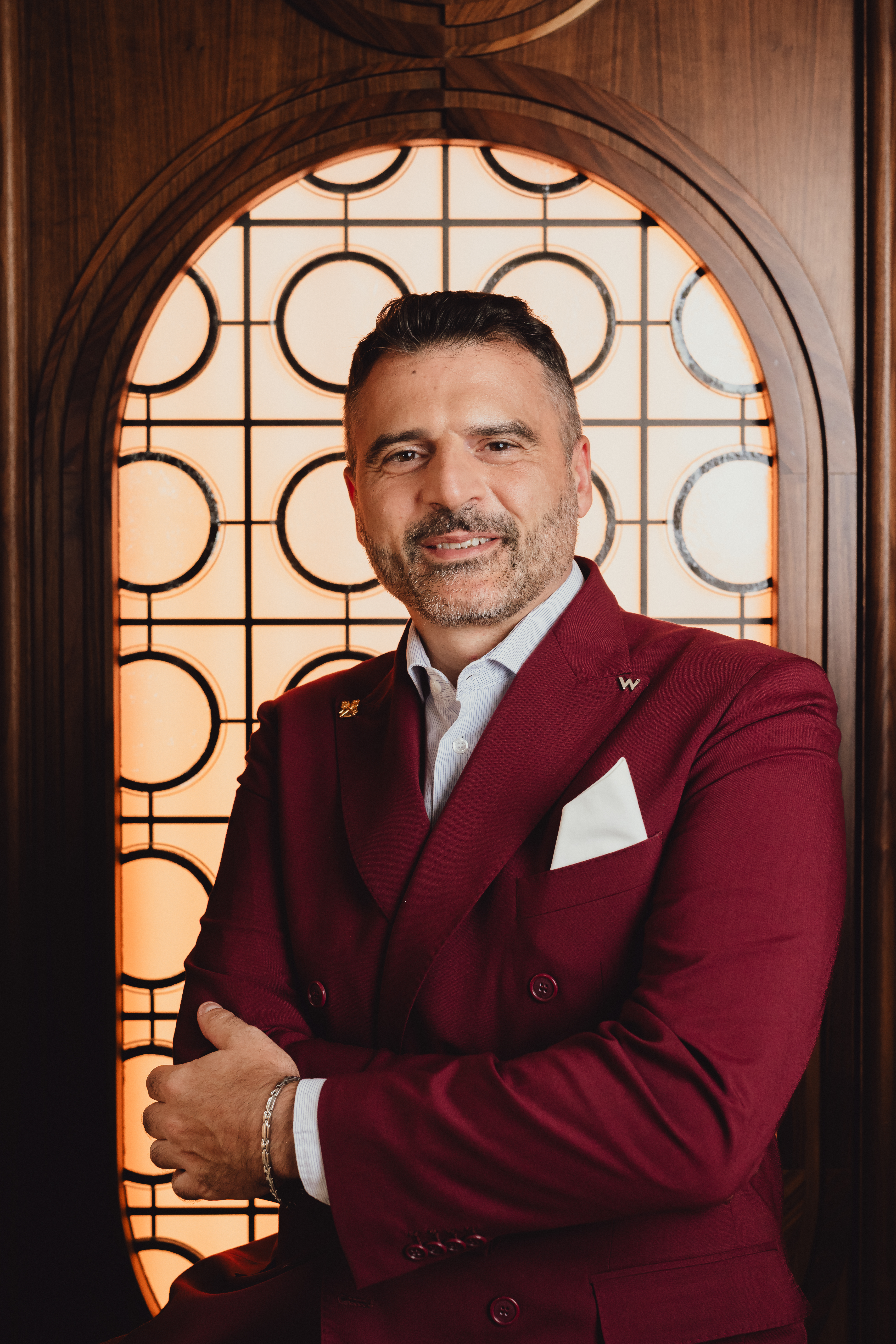Homem de meia-idade, barba grisalha e sorriso gentil, veste um terno vinho com camisa listrada e lenço branco no bolso, braços cruzados. Ele está em pé, em frente a uma janela decorativa de madeira escura com detalhes circulares e retangulares, iluminada por trás
