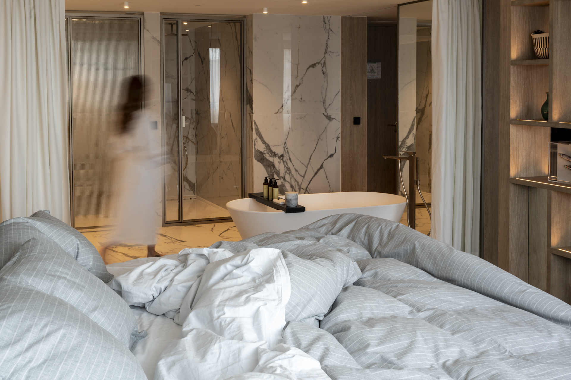 Quarto de hotel luxuoso com cama desfeita em primeiro plano e uma pessoa borrada em roupão branco caminhando em direção a uma banheira oval e um chuveiro de vidro ao fundo. As paredes são de mármore branco com veios escuros e há prateleiras de madeira iluminadas à direita