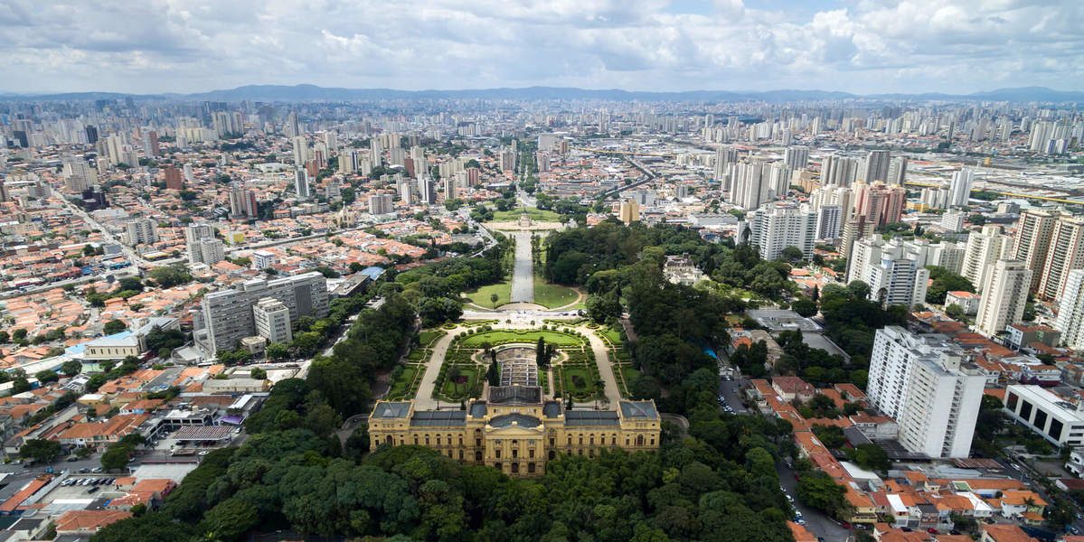 Vista aérea do Museu do Ipiranga em São Paulo, um edifício amarelo neoclássico cercado por jardins verdes e uma longa avenida com espelho dágua, em contraste com a vasta paisagem urbana de prédios altos e casas com telhados alaranjados sob um céu nublado