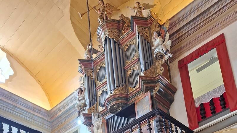 Órgão de tubos barroco em madeira escura, com detalhes dourados e vermelhos, adornado com esculturas de anjos tocando instrumentos, sob um teto abobadado claro e ao lado de uma janela com cortinas vermelhas