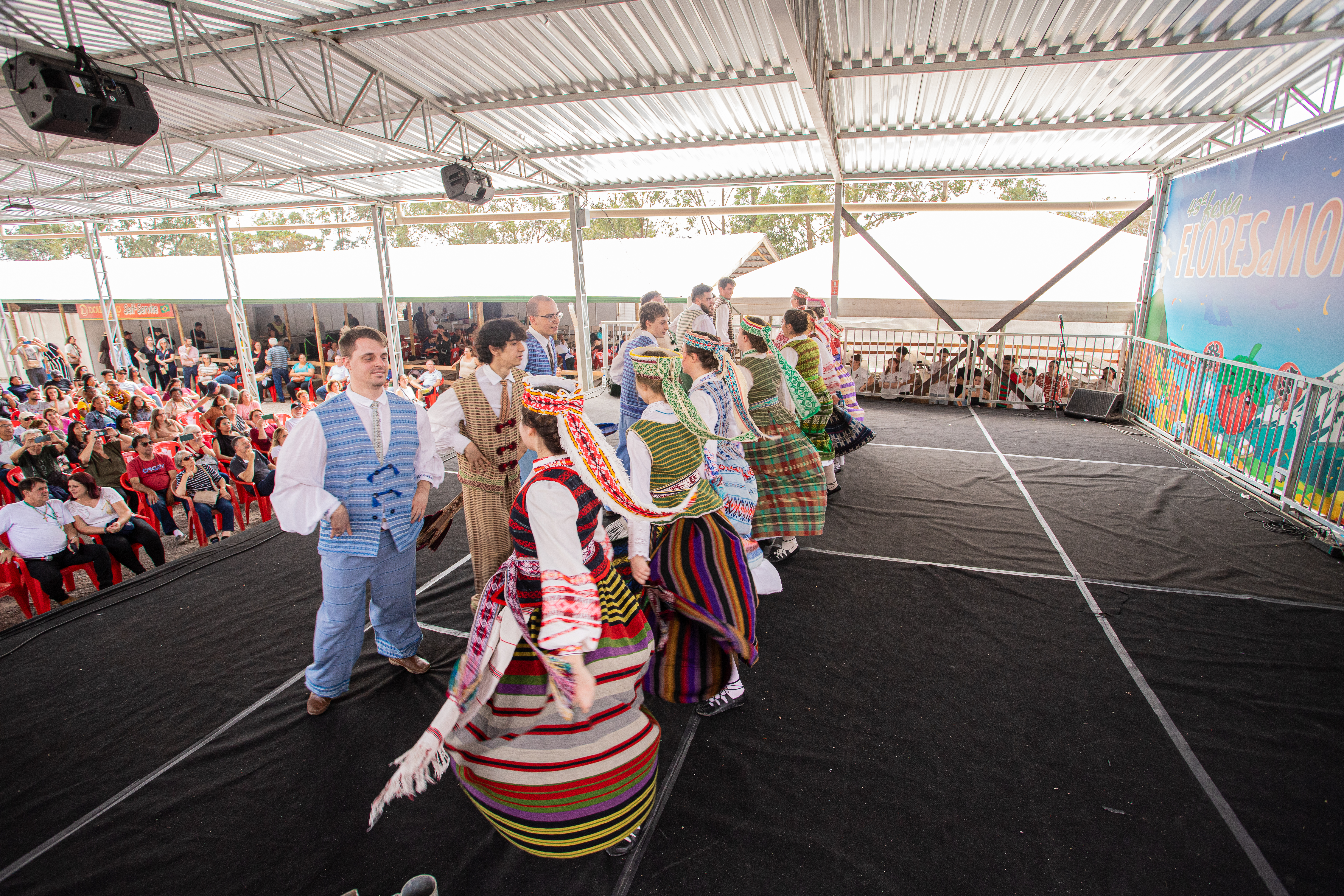 Grupo de dançarinos com trajes típicos coloridos, incluindo saias listradas e coletes, apresentando-se em um palco coberto. Um homem sorri para a câmera. Ao fundo, uma plateia sentada em cadeiras vermelhas e um banner azul com a frase FLORES MOR