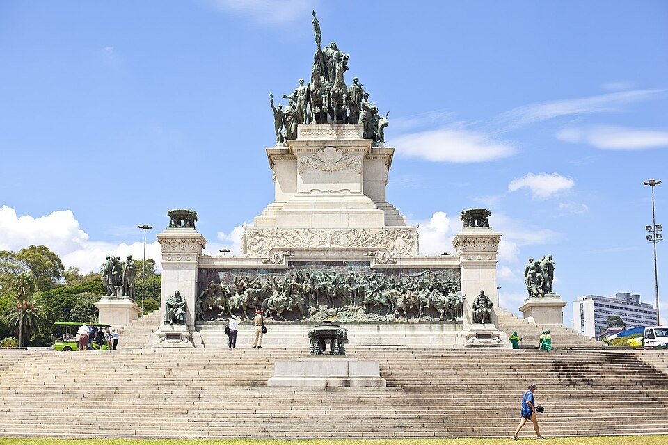 Monumento à Independência em São Paulo, com escadaria larga e pessoas pequenas subindo e descendo. O céu azul tem nuvens esparsas e árvores verdes ao fundo