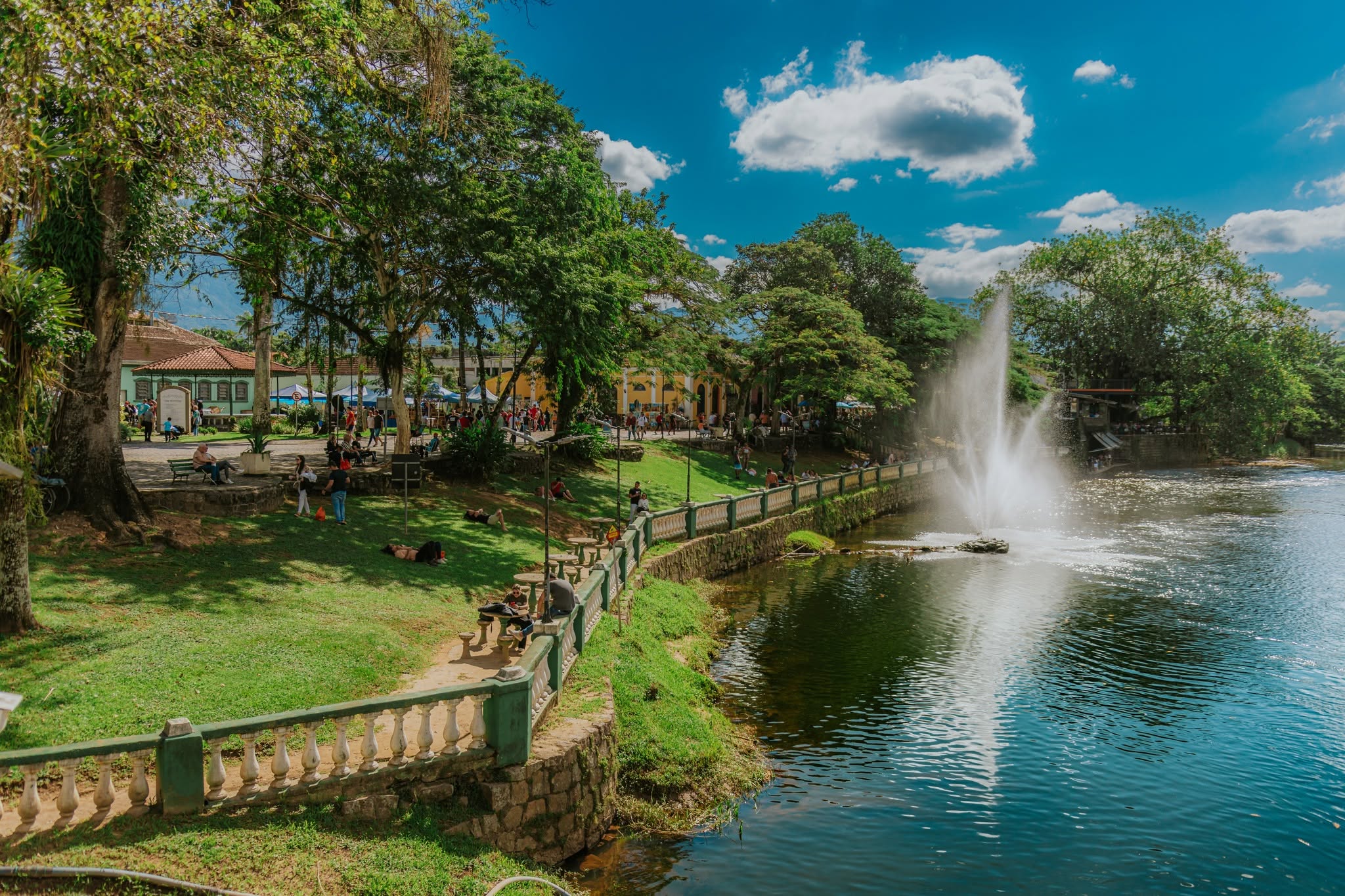 Parque urbano com gramado verde, árvores frondosas e pessoas relaxando. À direita, um lago com uma fonte jorrando água, ladeado por um muro de pedra e um corrimão branco e verde. Ao fundo, construções históricas e mais pessoas circulando sob um céu azul com nuvens brancas